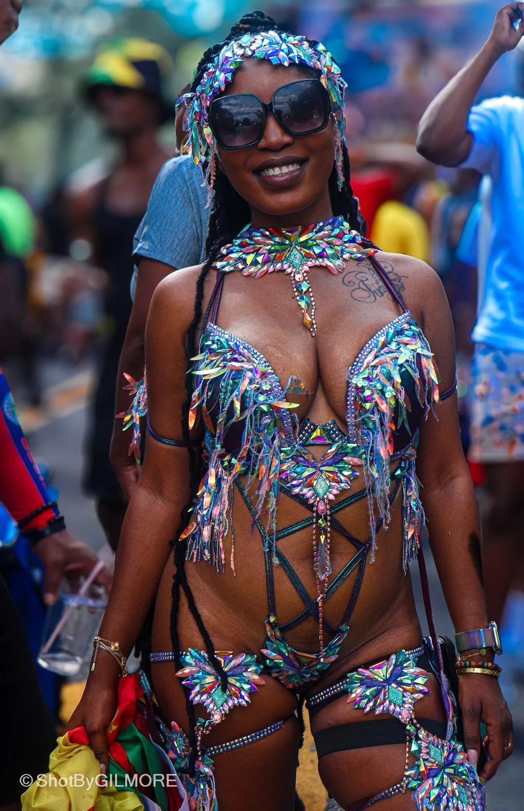 Woman smiling wearing a colorful, sparkling carnival costume with sunglasses, surrounded by other festival attendees.