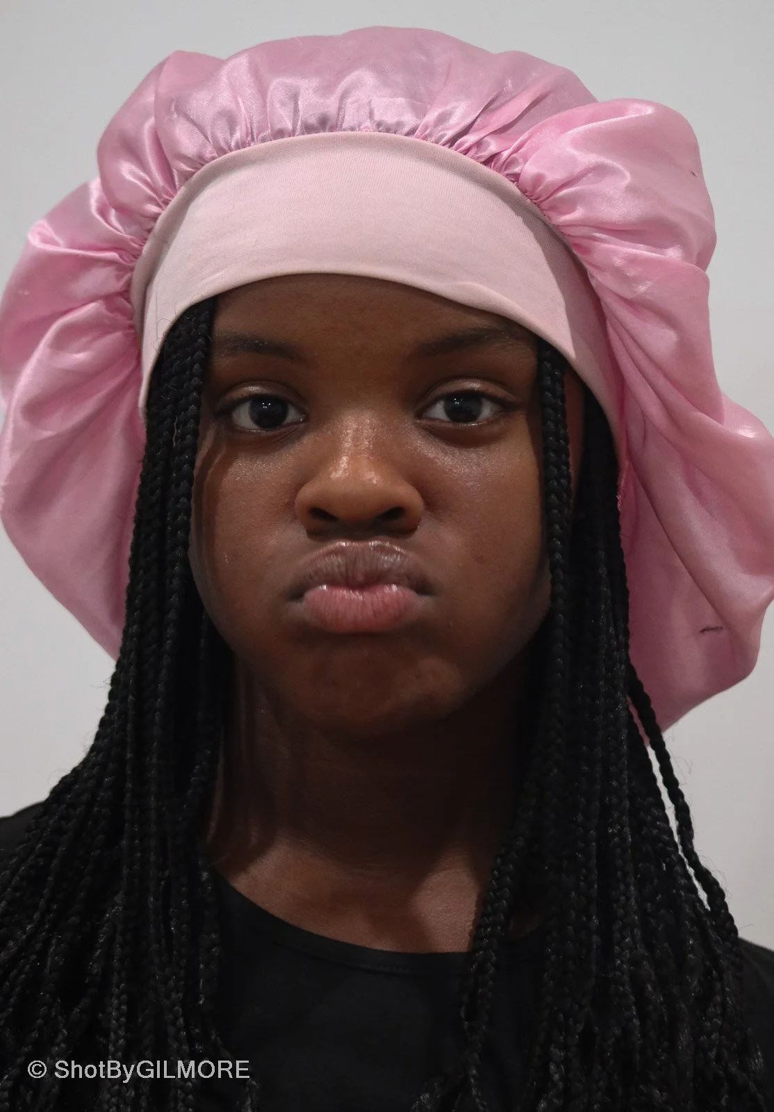 A young woman with dark skin and braided hair wearing a pink bonnet and matching satin headscarf, looking directly at the camera with a neutral expression.