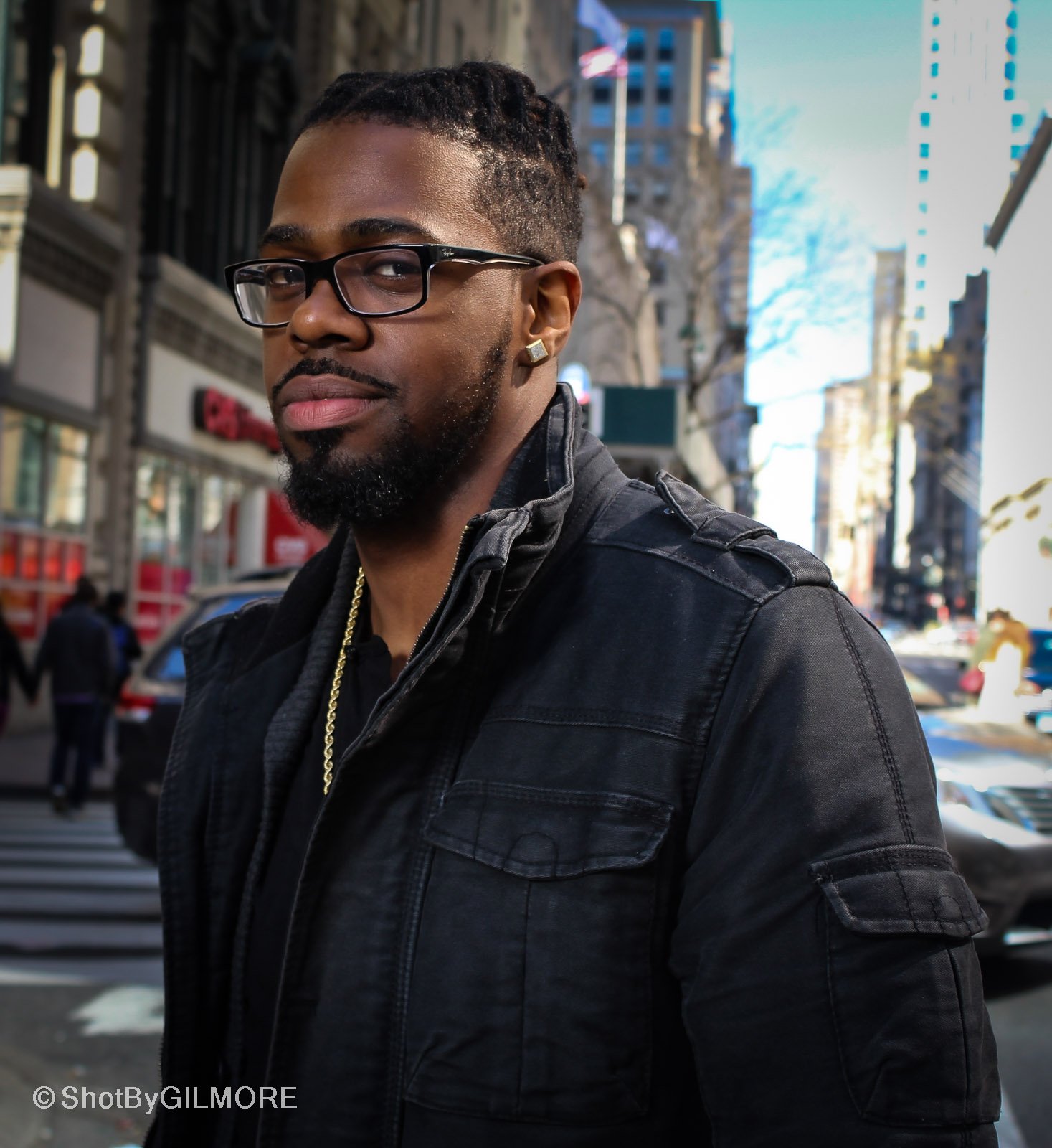 A man with glasses, earrings, and a beard poses on a city street with tall buildings, cars, and pedestrians in the background.