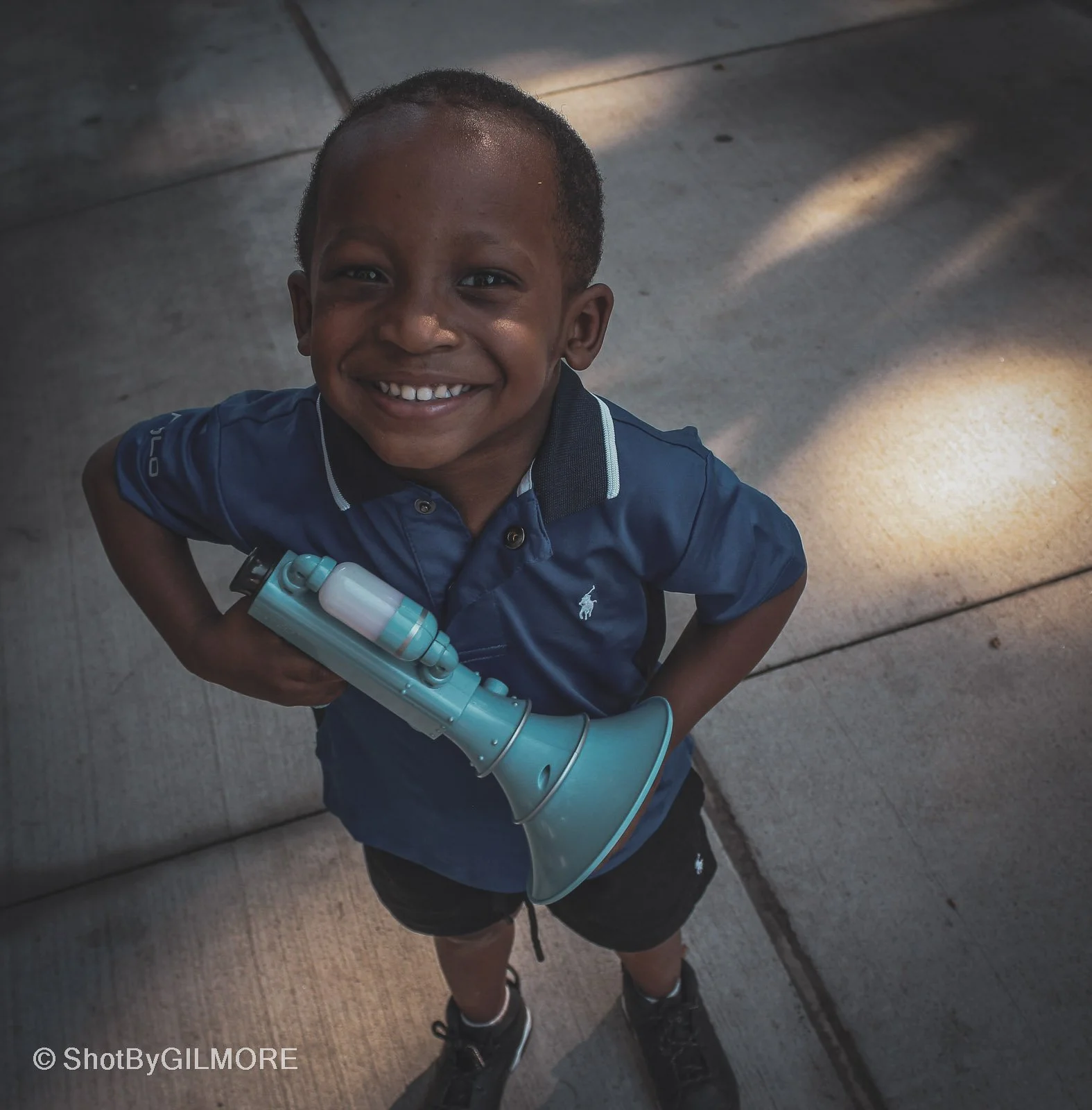 A smiling young boy in a blue polo shirt and shorts, holding a toy horn, looking up at the camera on a concrete sidewalk with sunlight patches.