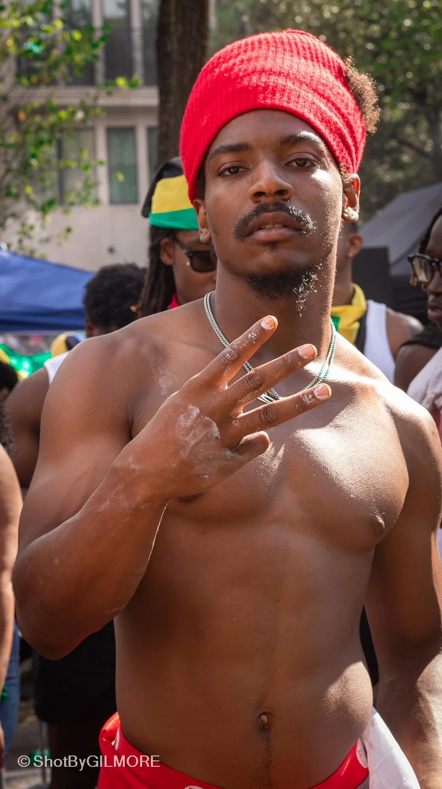 Young man with a red headwrap making a peace sign at a street event, with other people and trees in the background.