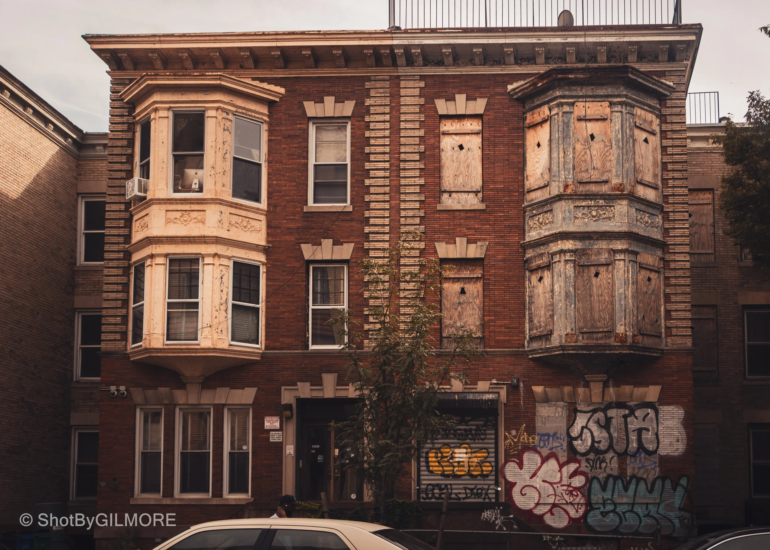 An old, abandoned four-story brick building with boarded-up windows and graffiti on the lower part of the facade. The building features decorative window frames and two prominent protruding bay windows with peeling paint. A small tree is in front, an