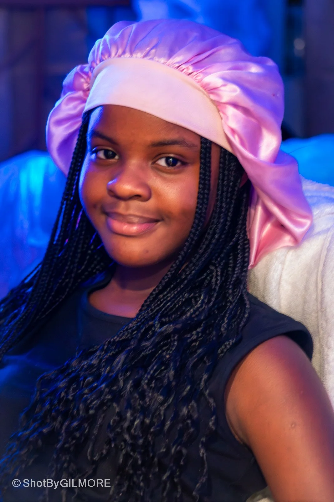 A young woman with braided hair wearing a pink satin bonnet and a black sleeveless shirt, smiling softly at the camera in a setting with blue ambient lighting.
