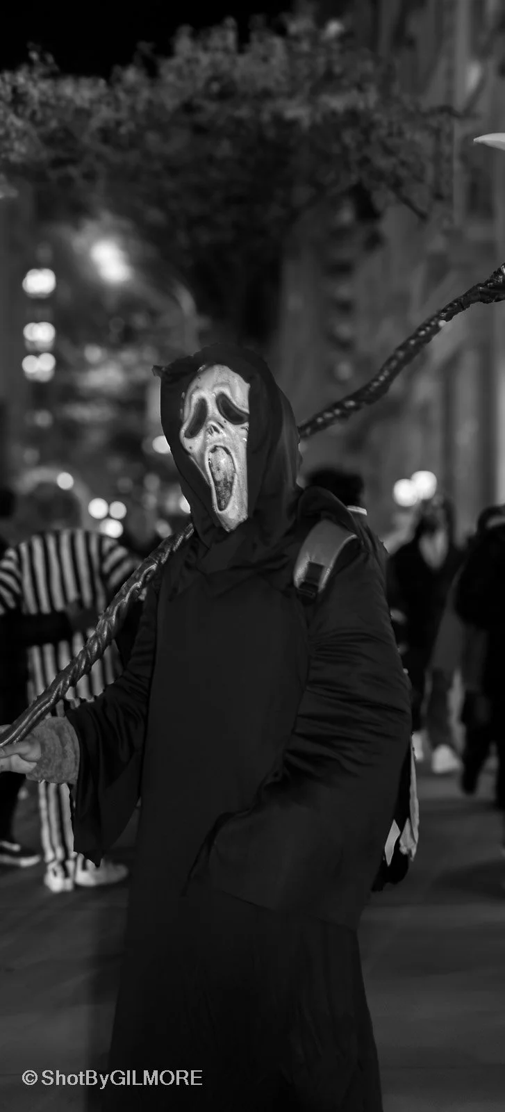 A person wearing a dark hoodie and a horror mask with a scream face, holding a large pole, in a black and white photo at night with blurred lights and people in the background.