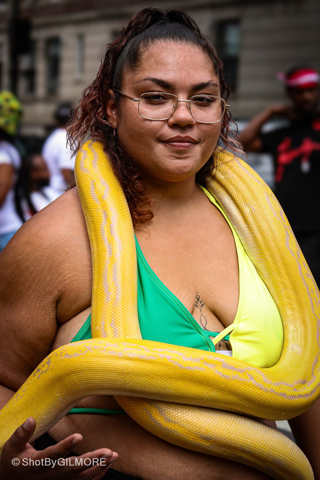 A woman wearing glasses and a colorful top with a yellow and gold snake draped over her shoulders and arms. She is outdoors, with other people and a building in the background.