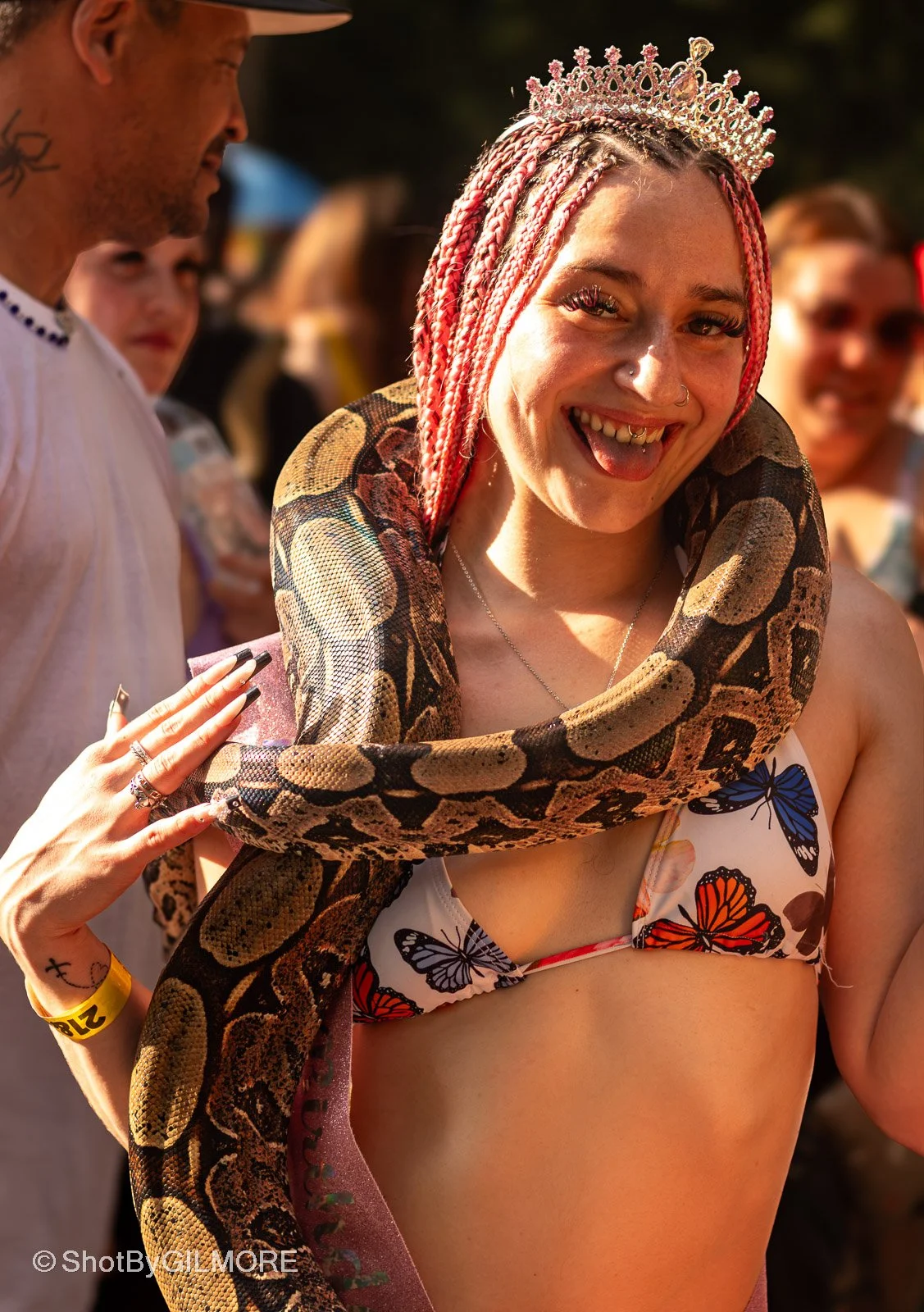 Woman with pink braids, wearing a crown and butterfly-printed bikini top, smiling with a large snake draped around her shoulders at an outdoor event.