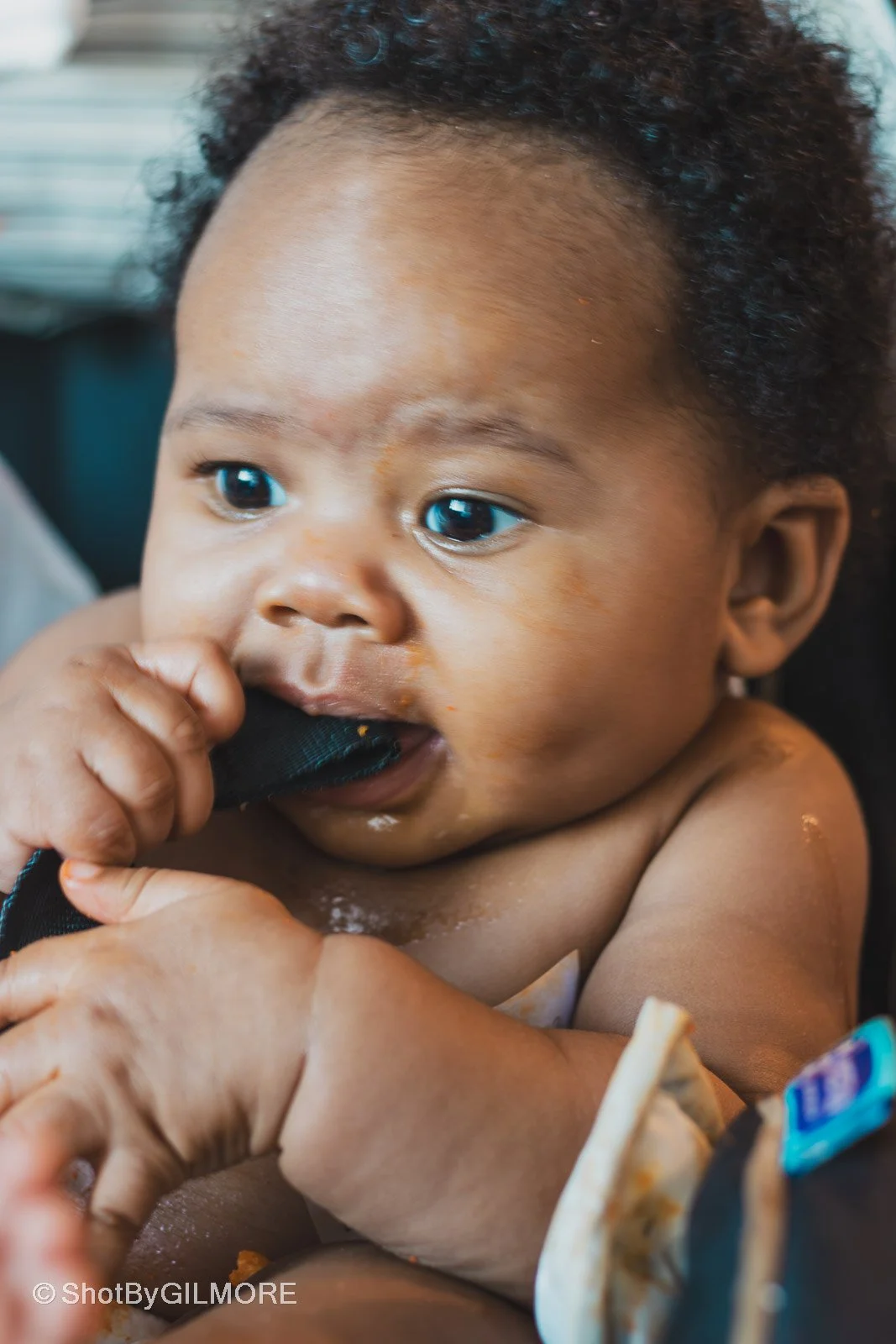 Close-up of a young child with dark skin and curly hair, chewing on a black strap, with food on their face and arms.