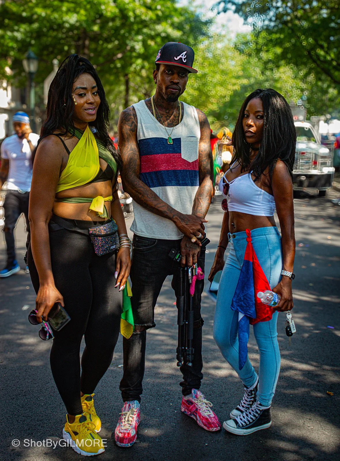 Three young adults, two women and one man, standing outdoors on a sunny day with trees in the background. They are dressed in casual, urban clothing and appear to be at a public event or gathering.