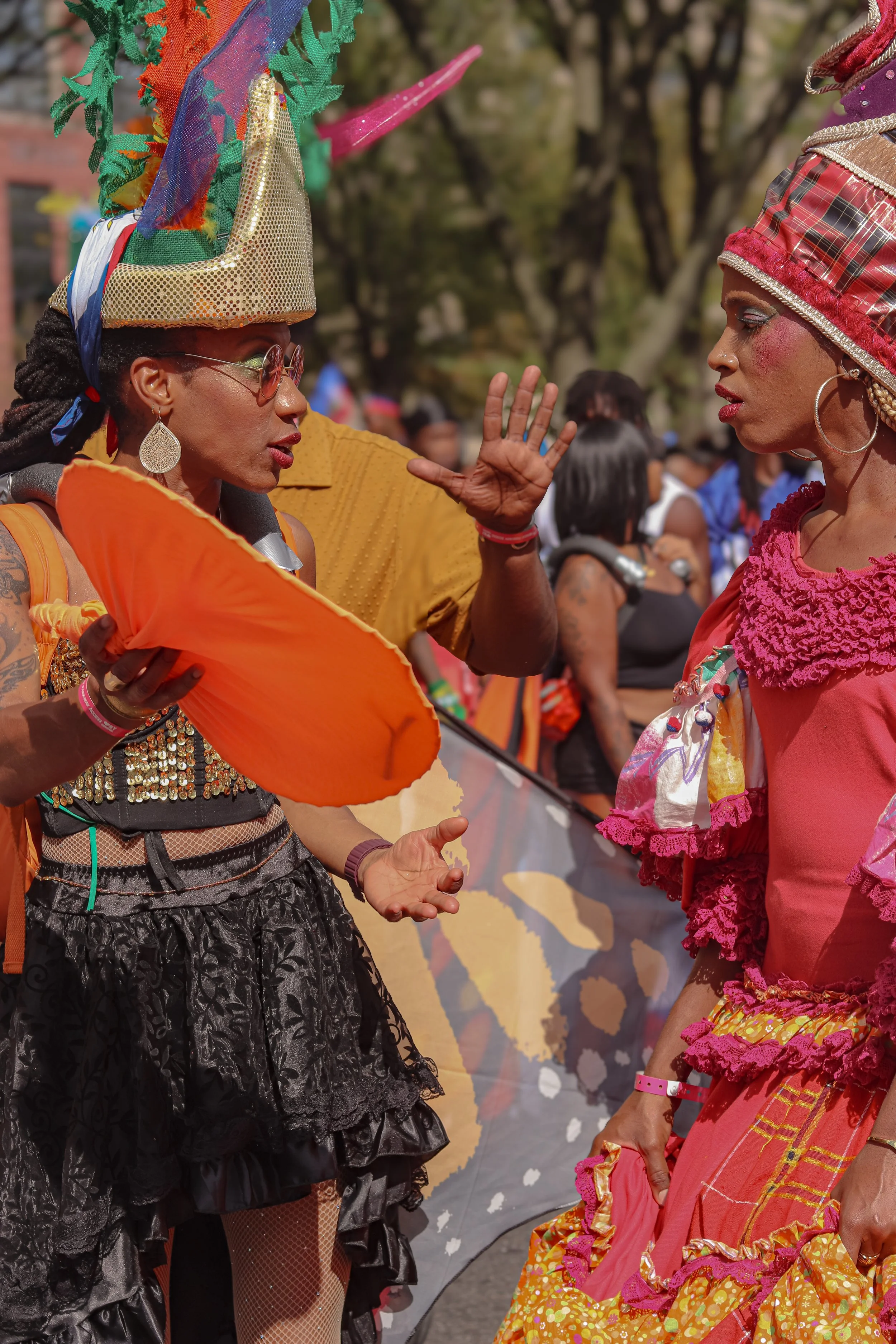 Two women in colorful costumes and headpieces are talking at a parade or festival, with other people in the background.