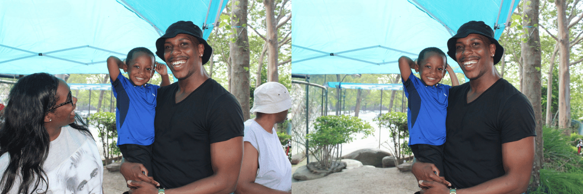 A man holding a smiling young girl on his shoulder at a park or zoo, with two women standing nearby, under a blue canopy, surrounded by trees and plants.