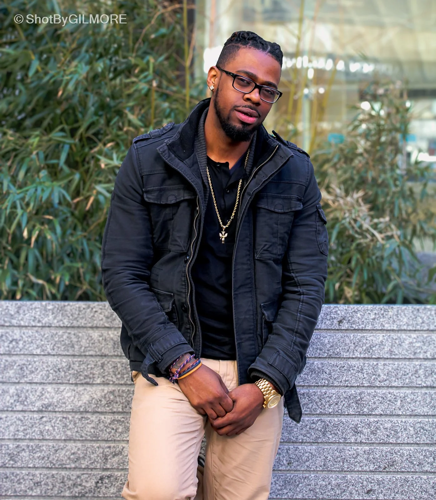 A young man with glasses, a beard, and braided hair standing outdoors in front of green bushes, wearing a black jacket, black shirt, beige pants, and gold jewelry.