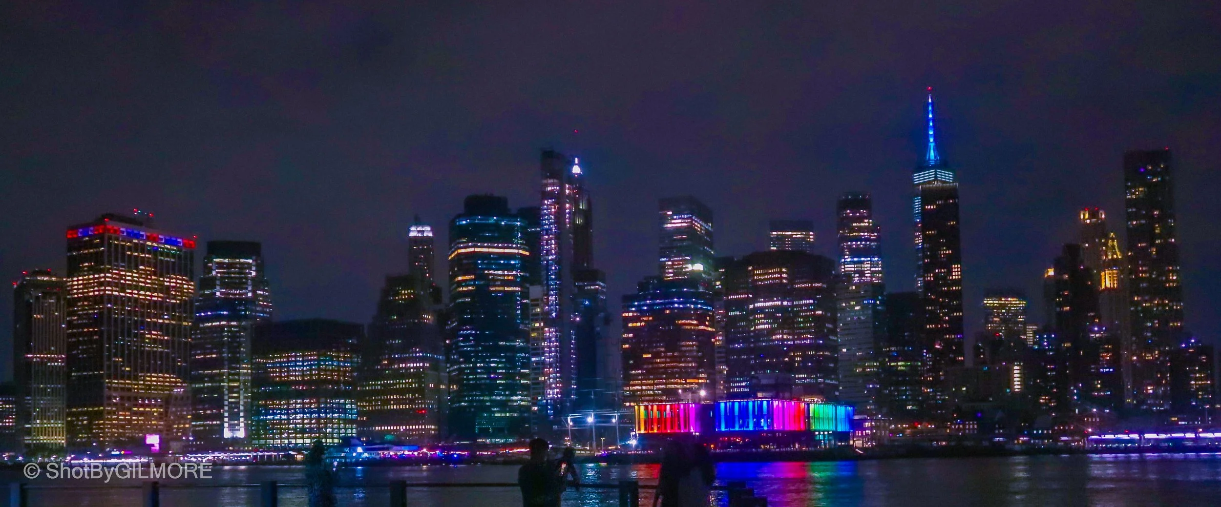 Night view of a city skyline with illuminated skyscrapers and a water reflection. The buildings are lit with colorful lights, including blue, purple, red, and yellow, with one building topped with blue lights.
