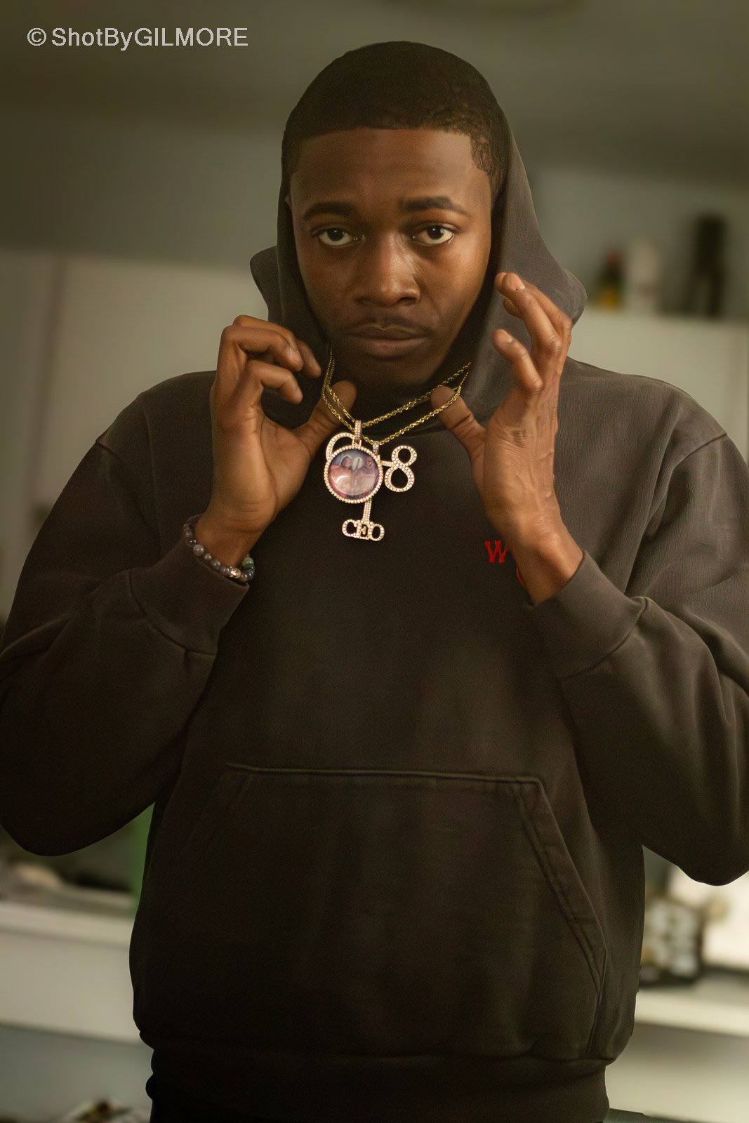 A young man with dark skin wearing a black hoodie, adjusting two gold necklaces with large pendants, in an indoor setting.