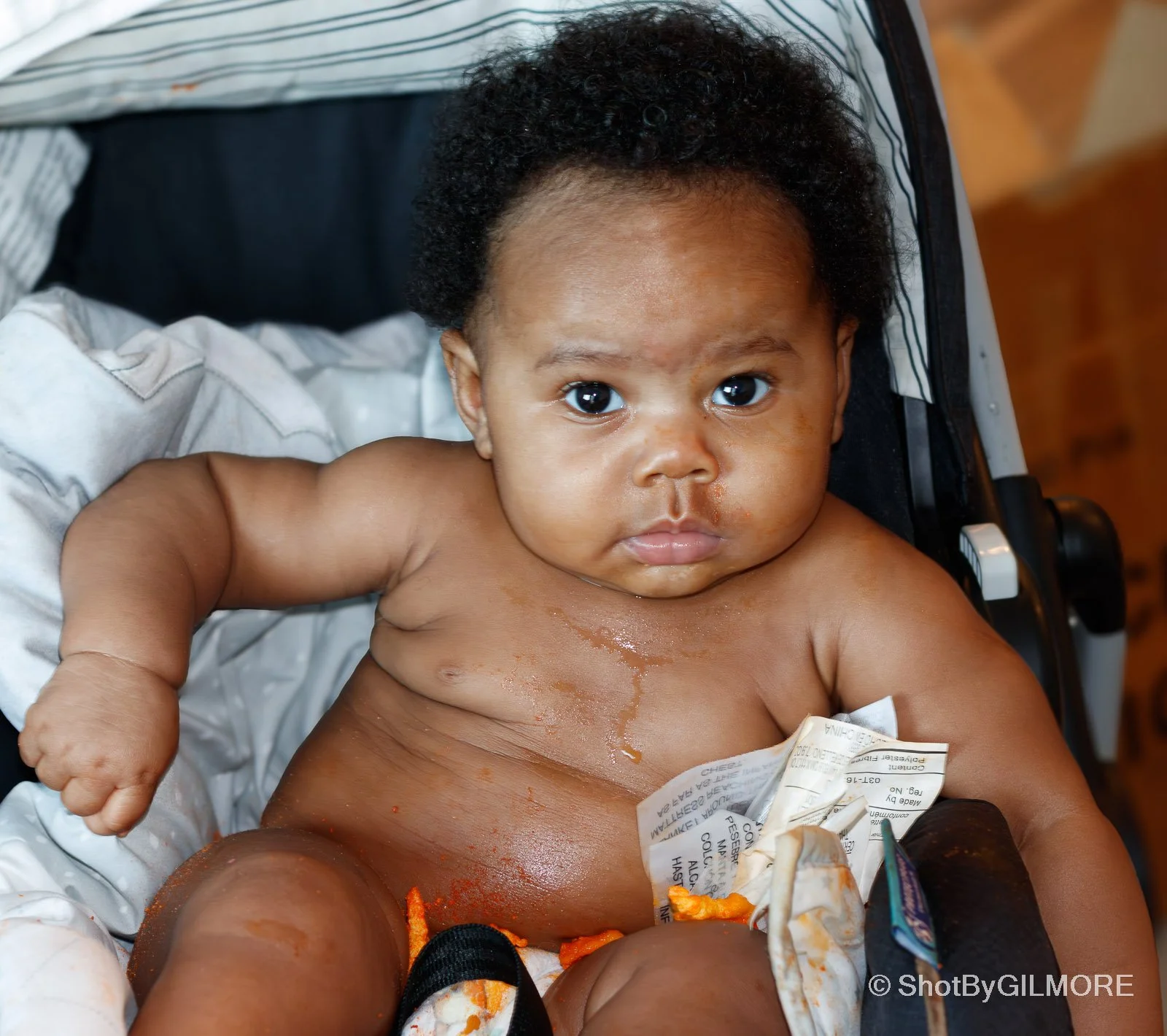 A shirtless baby sitting in a stroller with food mess on face and body, holding a torn package, looking directly at the camera.