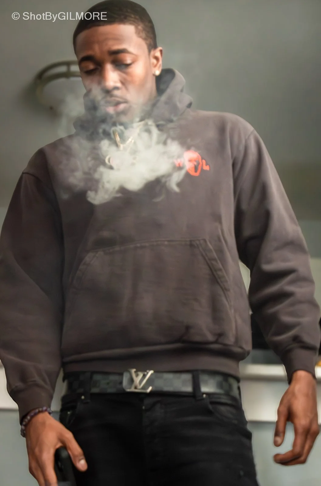 A young man in a black hoodie with red logo, black Louis Vuitton belt, and dark jeans, standing indoors with smoke around his face, holding a phone in his left hand.