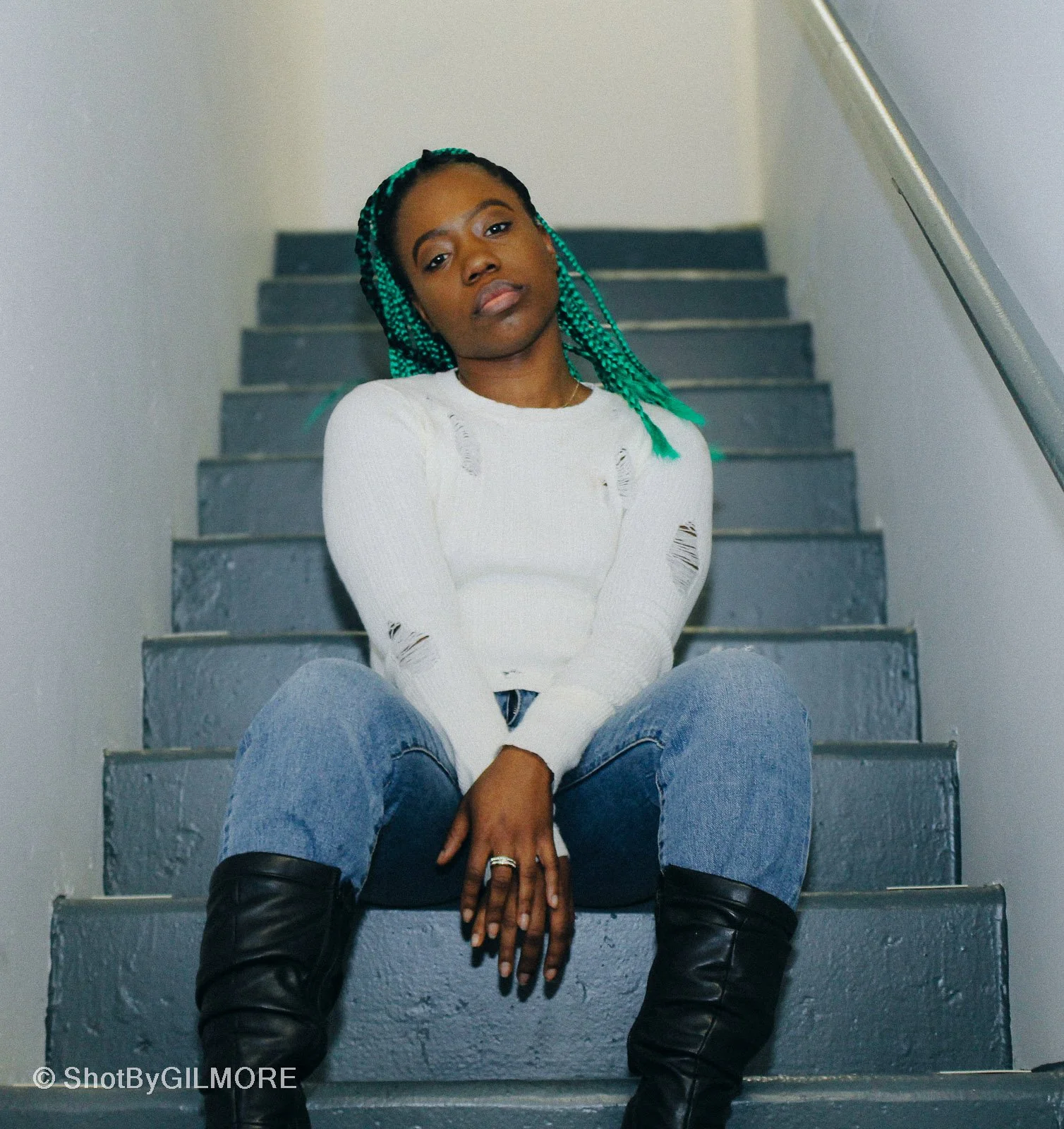 A woman with teal braids sitting on gray stairs, wearing a white distressed sweater, blue jeans, and black knee-high boots.