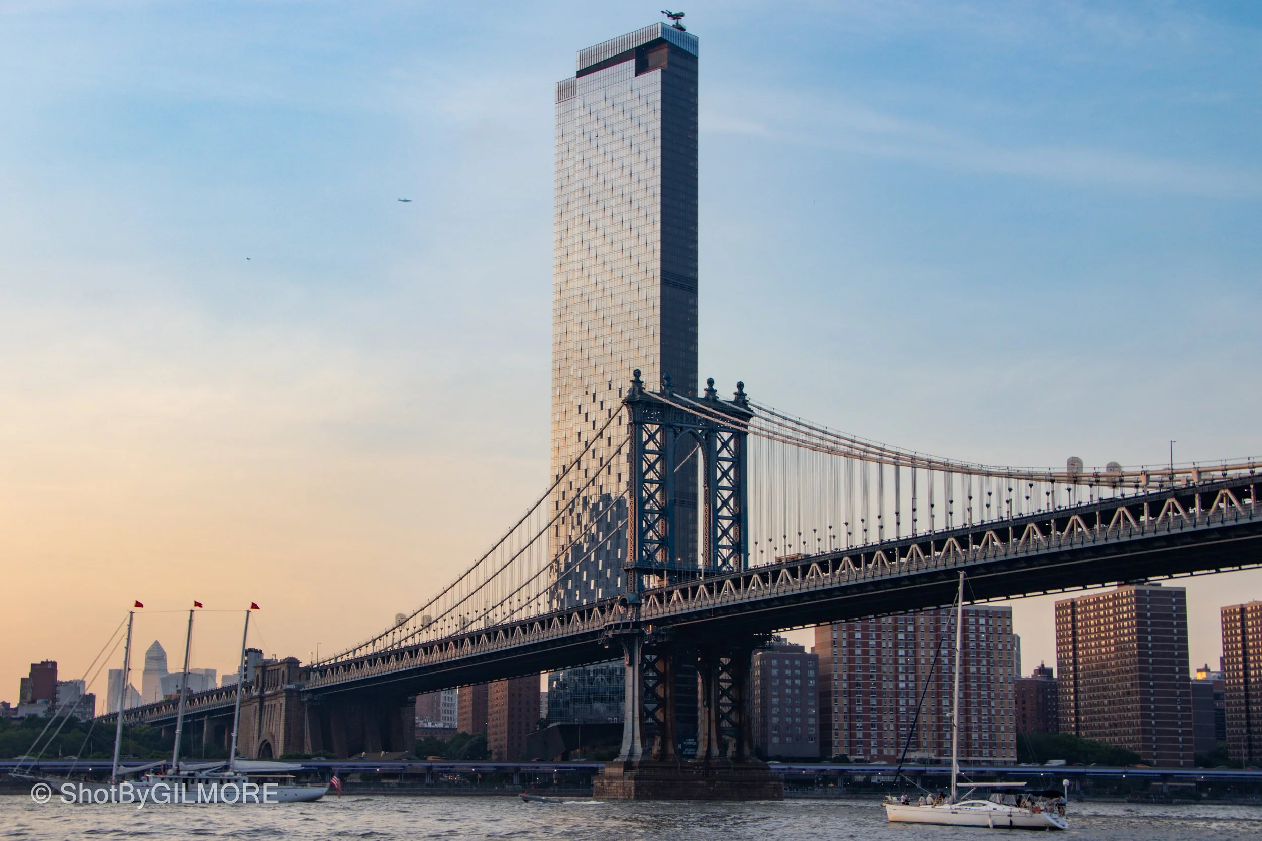 View of the Brooklyn Bridge with sailboats on the water and the Manhattan skyline in the background, including a tall skyscraper.