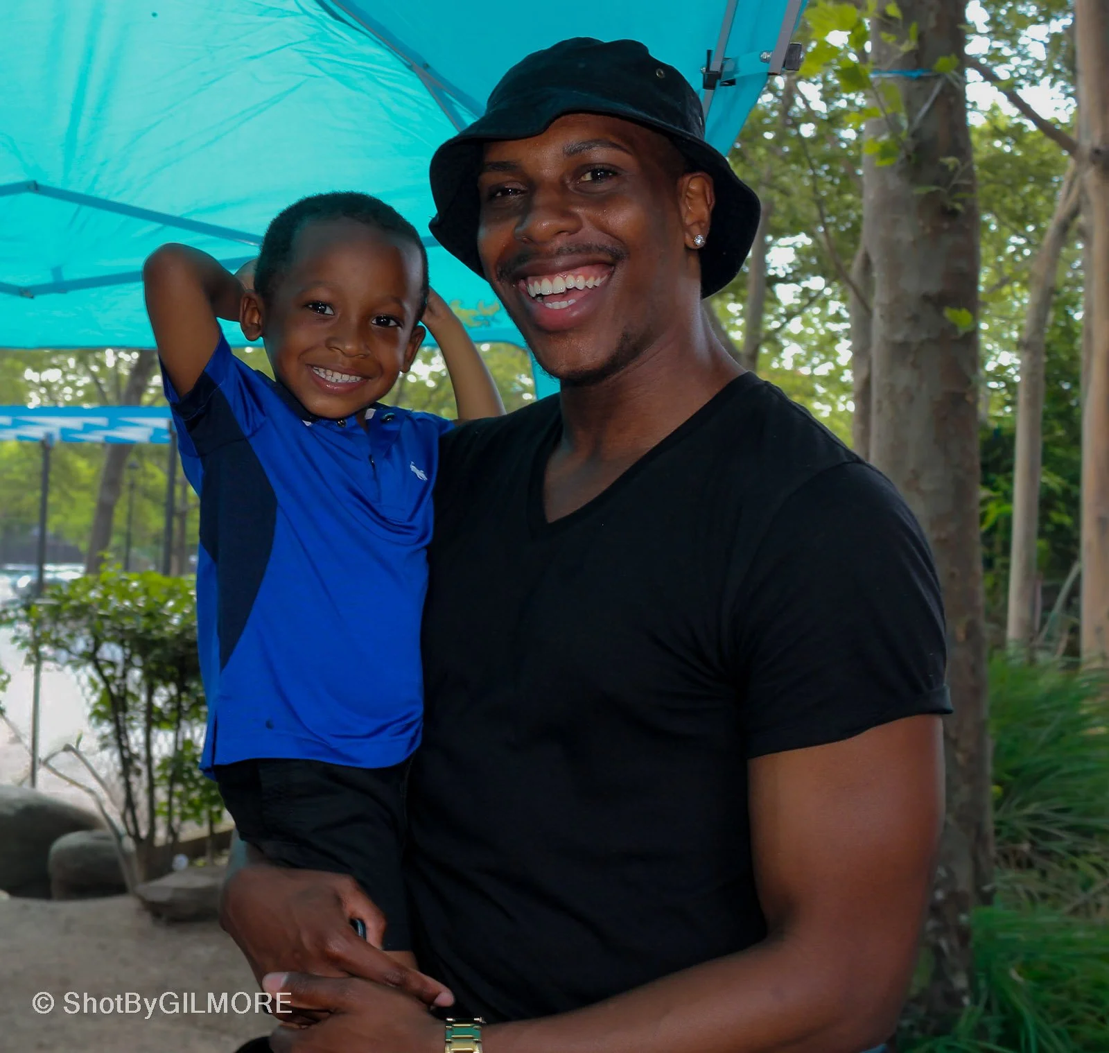 A man holding a happy young boy outdoors under a blue canopy with trees and bushes in the background, both smiling at the camera.