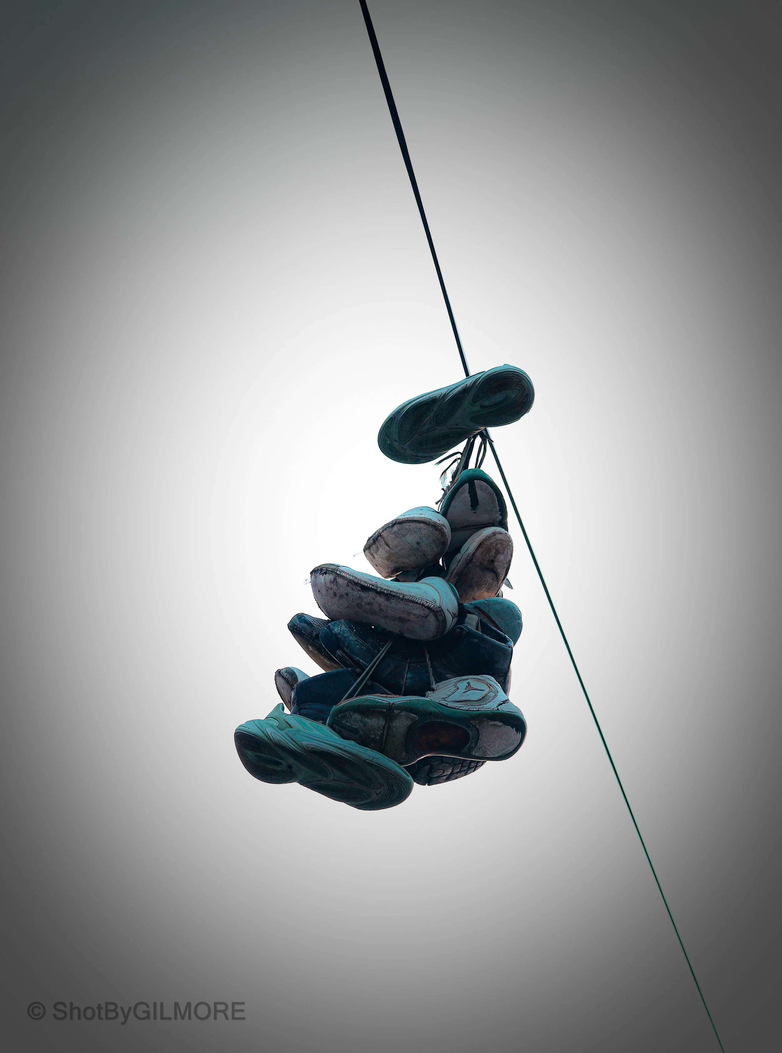 A cluster of various shoes hanging from a rope against a cloudy sky.