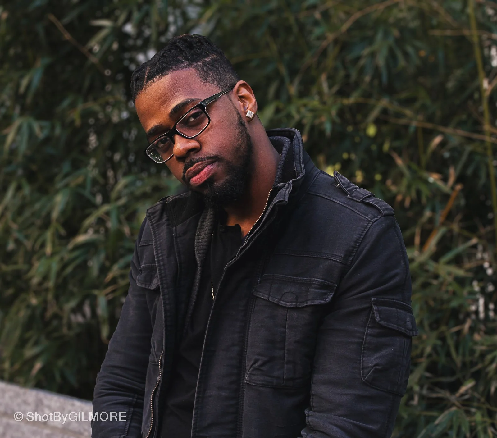 A young man wearing glasses and a black jacket, posing outdoors with a background of green foliage.