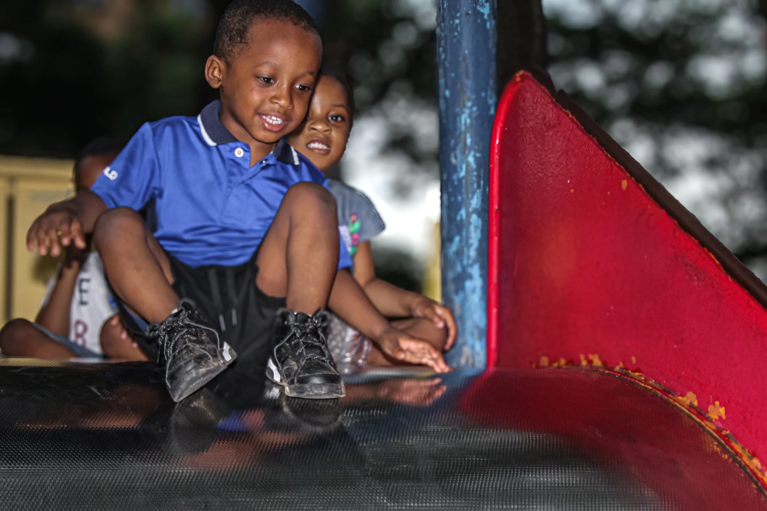 Children playing on a playground slide, with a boy in front wearing a blue shirt and black shorts, smiling, and a girl in the background.