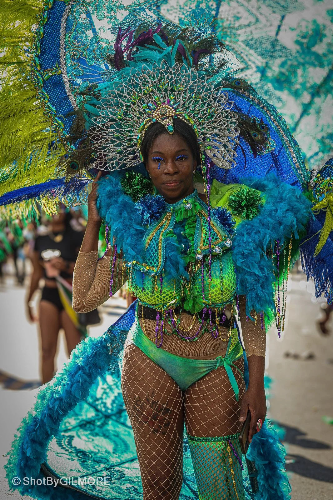 A woman dressed in a vibrant, elaborate costume featuring bright blue, green, and purple feathers, beads, and intricate designs, participating in a carnival parade.