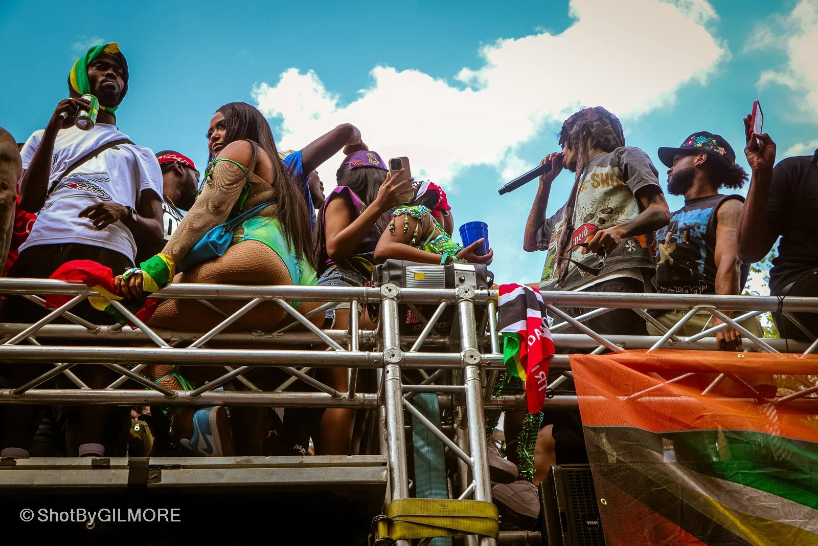 A group of people on a raised platform, some wearing colorful clothing and accessories, at an outdoor event under a partly cloudy sky.