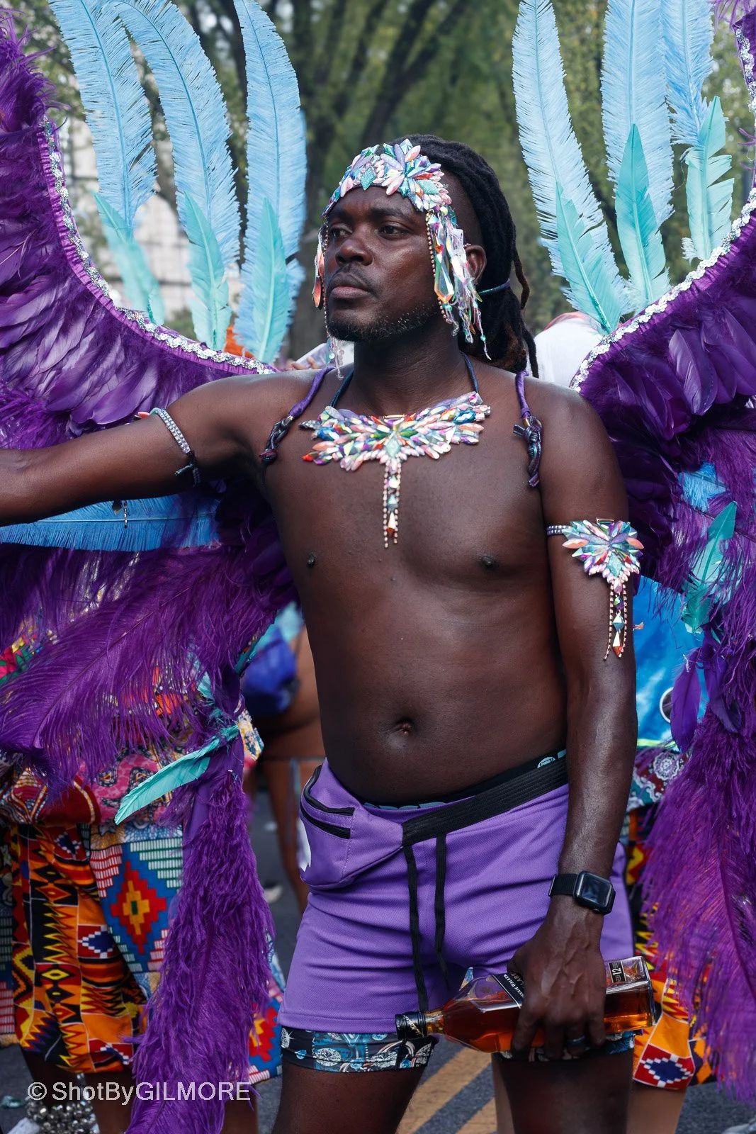 A man in colorful carnival attire, with large purple feathers on his back, wearing a jeweled headpiece and necklace, holding a bottle of alcohol at a parade or festival.
