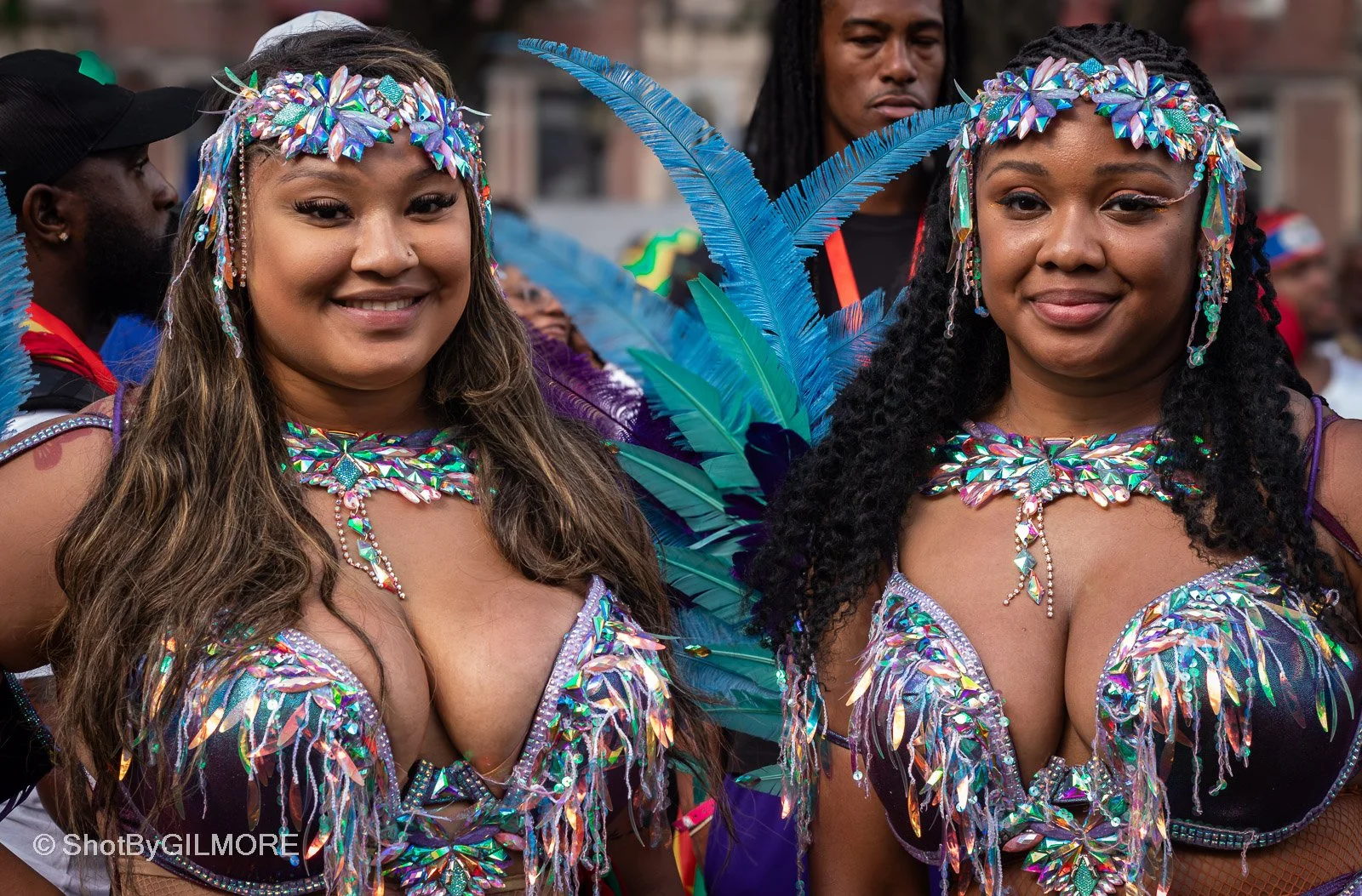 Two women dressed in colorful carnival costumes adorned with iridescent jewels and feathers, smiling at a festival or parade.