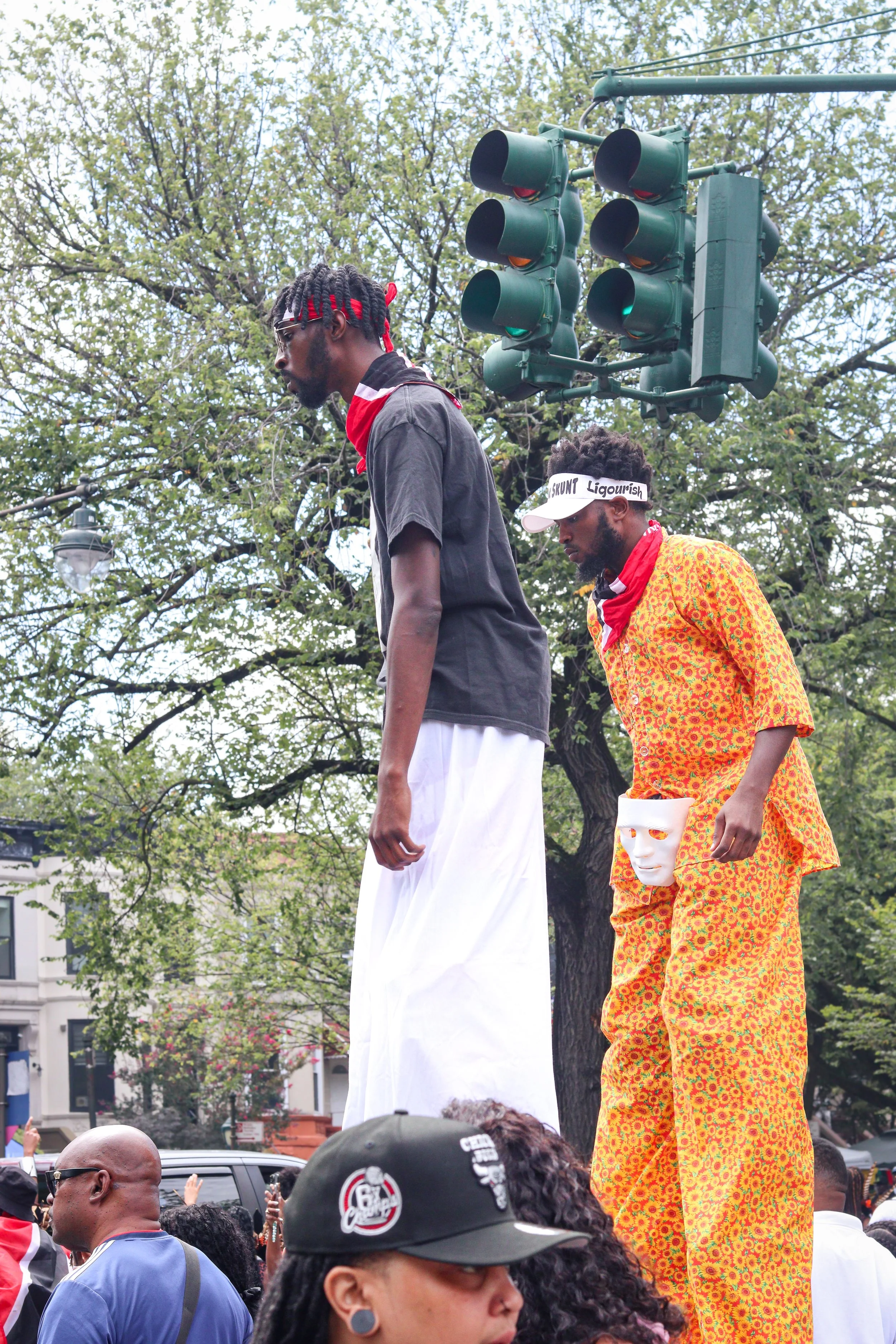 Two tall mannequins stand on a raised platform during a protest or event, dressed in casual clothing with one wearing a black shirt and long white skirt, and the other in a brightly patterned orange outfit holding a white mask, with a crowd of people