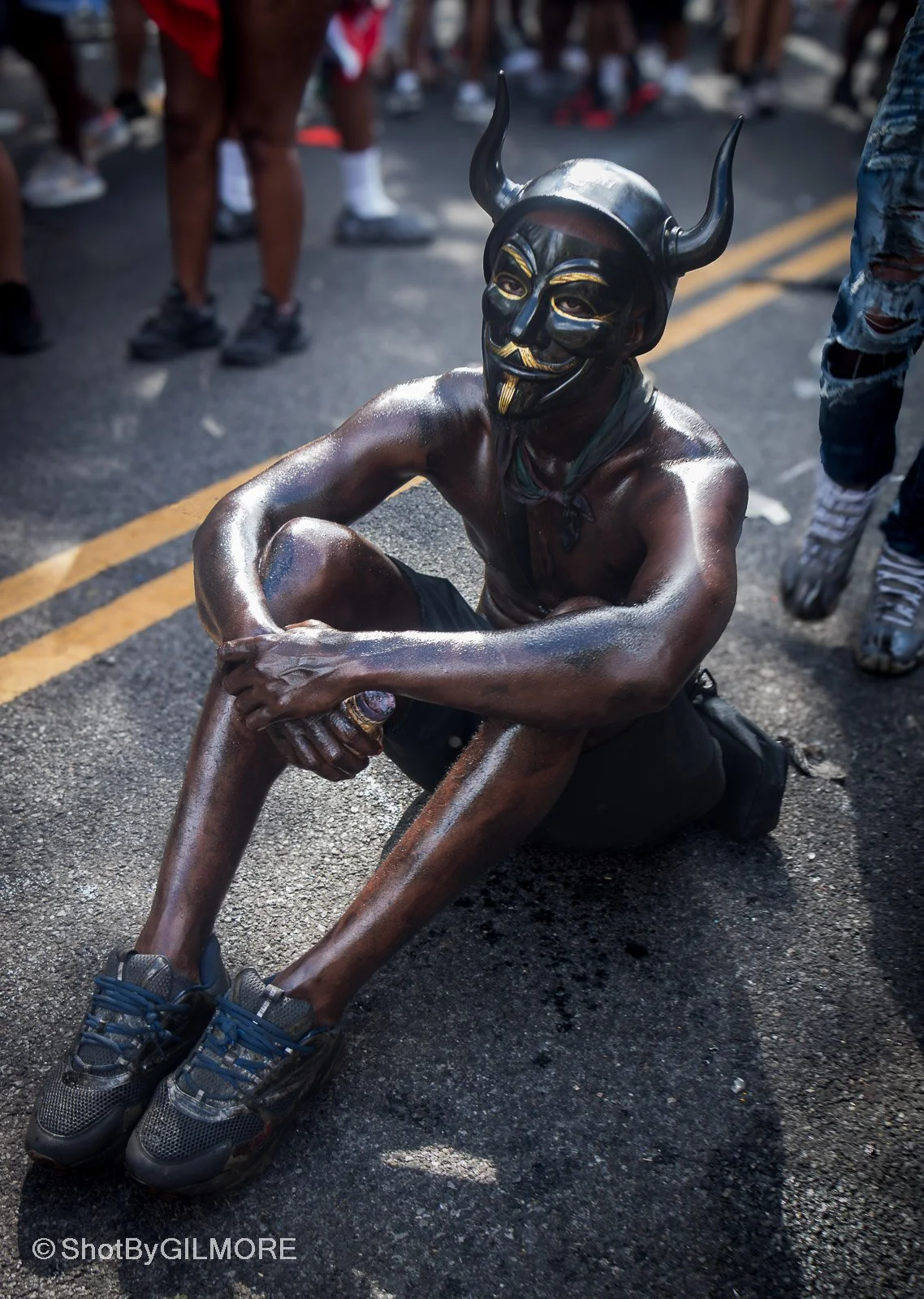 Person wearing a black mask with horns and gold details, shirtless and sitting on pavement during street event with other people in the background.