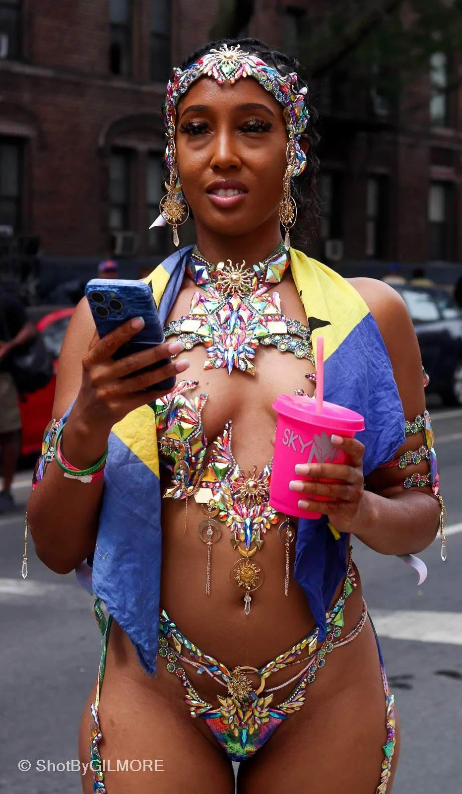 Woman dressed in colorful, elaborate costume with jewelry, holding a smartphone in one hand and a pink cup with a straw in the other, standing outdoors in an urban setting.