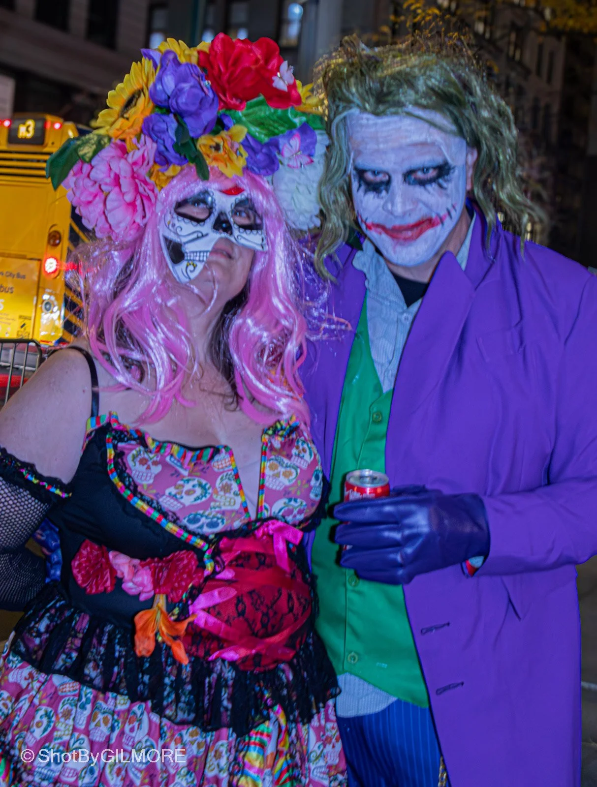 Two people dressed as characters from the Joker and Día de los Muertos, standing close together on a city street at night, with yellow bus in background.