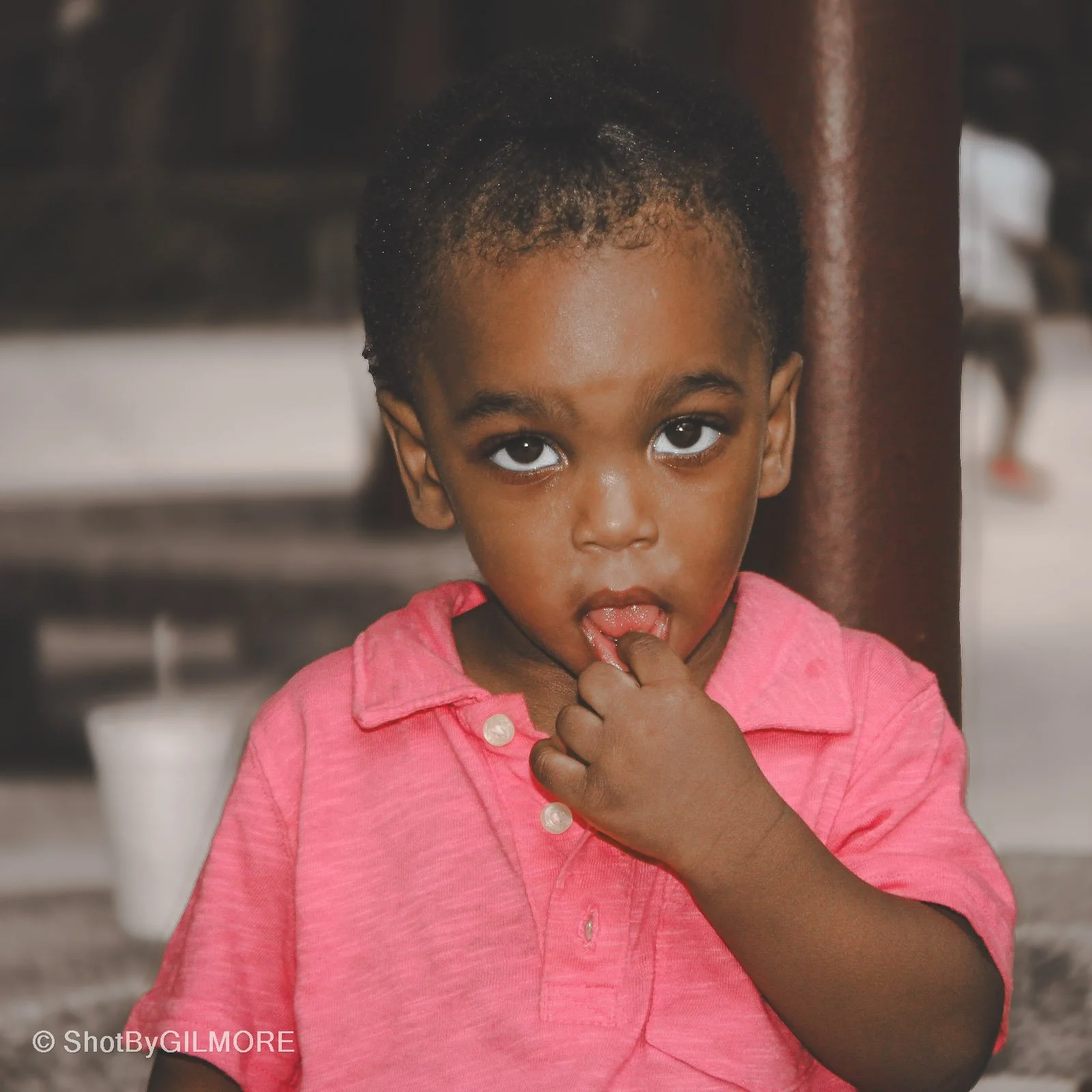 Young girl with dark hair, wearing a pink shirt, looking at the camera with her finger in her mouth.
