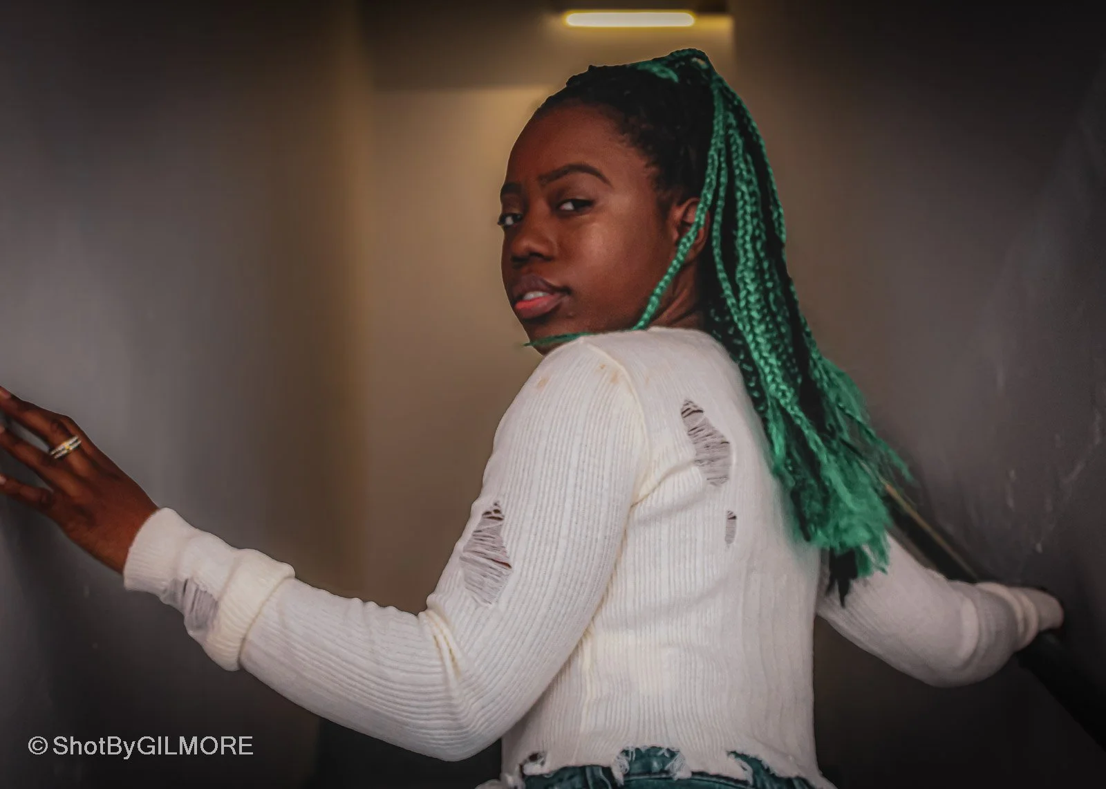 A young woman with green box braids wearing a ripped white sweater and standing in a dimly lit corridor.