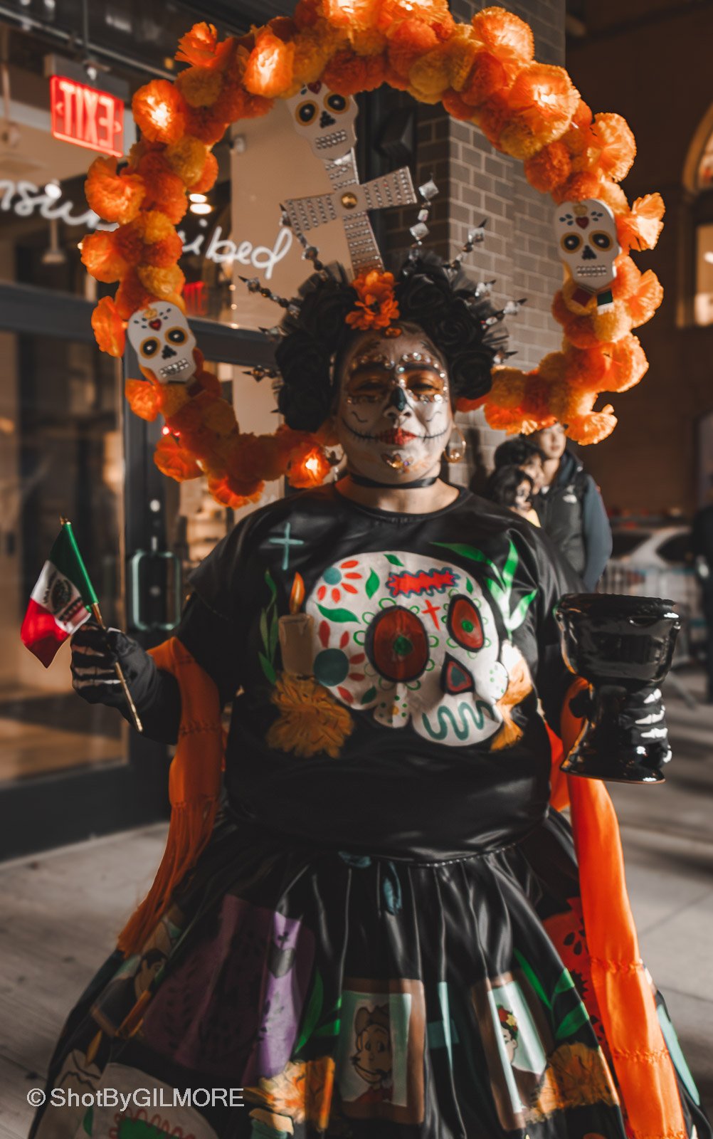 A person dressed in Día de los Muertos (Day of the Dead) themed costume with face paint resembling a decorated skeleton, wearing an elaborate headdress with skull decorations and orange flowers, holding a small Mexican flag and a ceramic bowl, standi