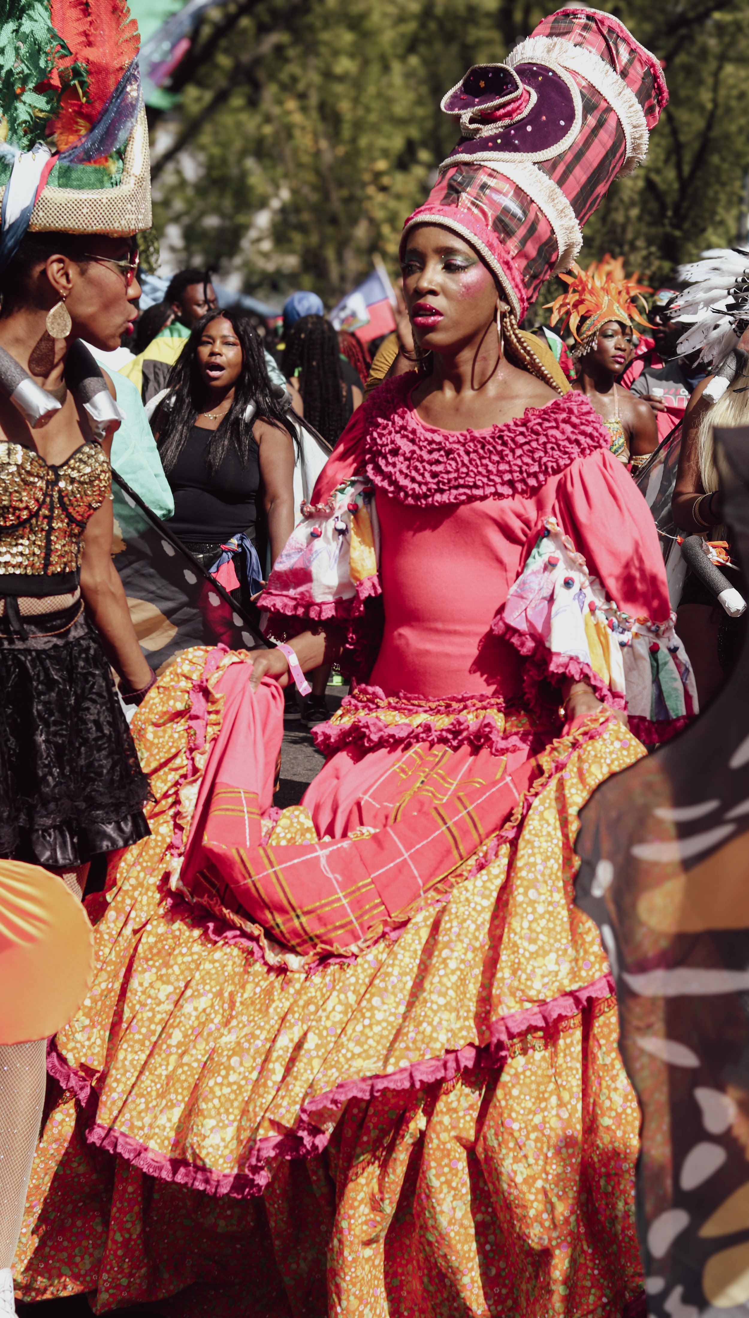 A woman dressed in a colorful, elaborate costume wearing a tall hat, participating in a parade or festival, surrounded by other people in festive attire.