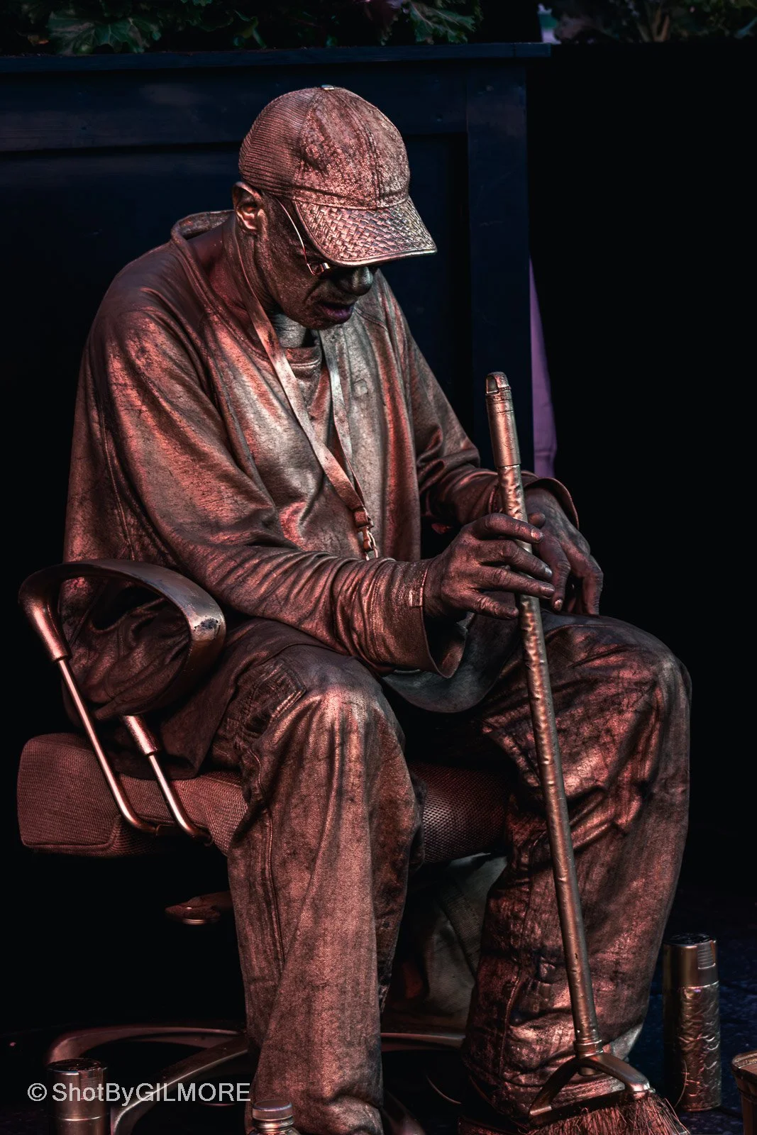 A person sitting on a chair, holding a metallic stick, wearing a cap, and clothes with a metallic or copper-colored look. The person appears to be performing or preparing for a performance.