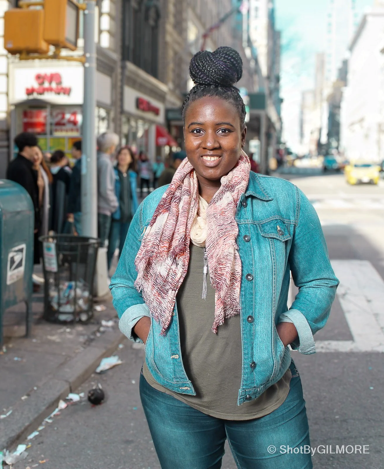A woman standing on a city street with her hands in her pockets, smiling at the camera. She has her hair in a bun, wearing a denim jacket and a patterned scarf. There are people, a trash can, and a storefront in the background.