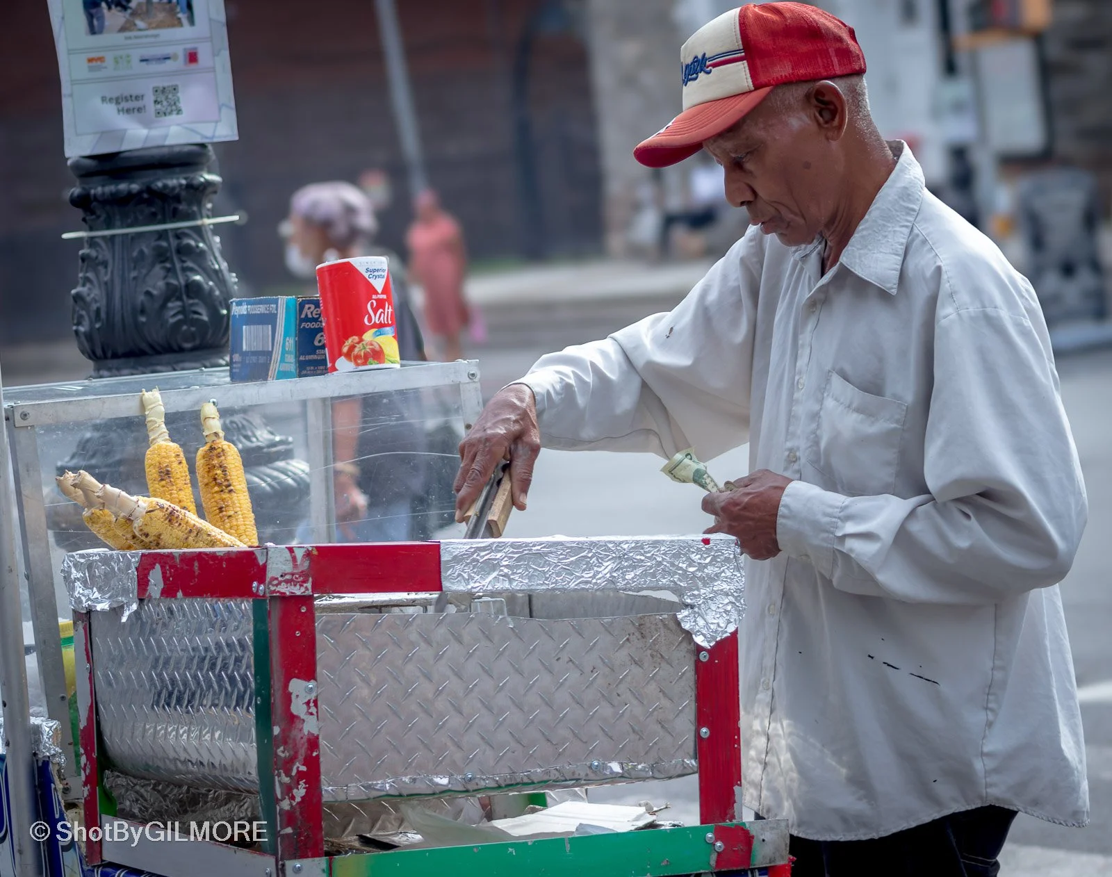 An elderly man in a white shirt and red cap stands at a street corn stand, preparing grilled corn on the cob with aluminum foil wrapping on the cart. There are salt and other condiments nearby, with a QR code sign above.