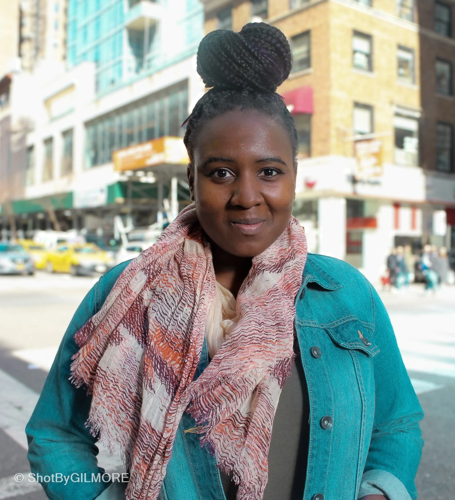 A woman with dark skin and braids styled in a bun, smiling while standing outdoors on a city street with buildings and cars in the background.
