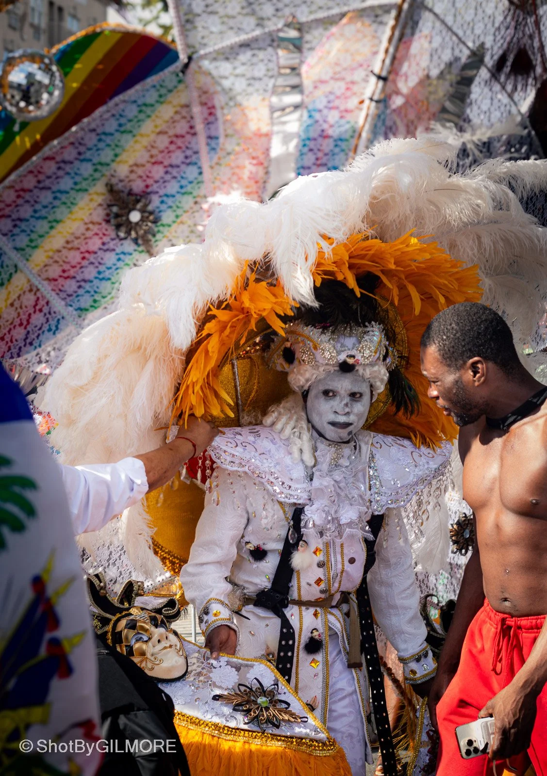 Person dressed in elaborate traditional costume with large feathered headdress and face paint surrounded by people at a cultural event.