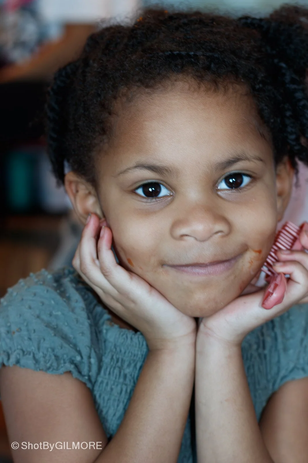 Close-up of a young girl with dark curly hair smiling, resting her face in her hands, and looking at the camera.