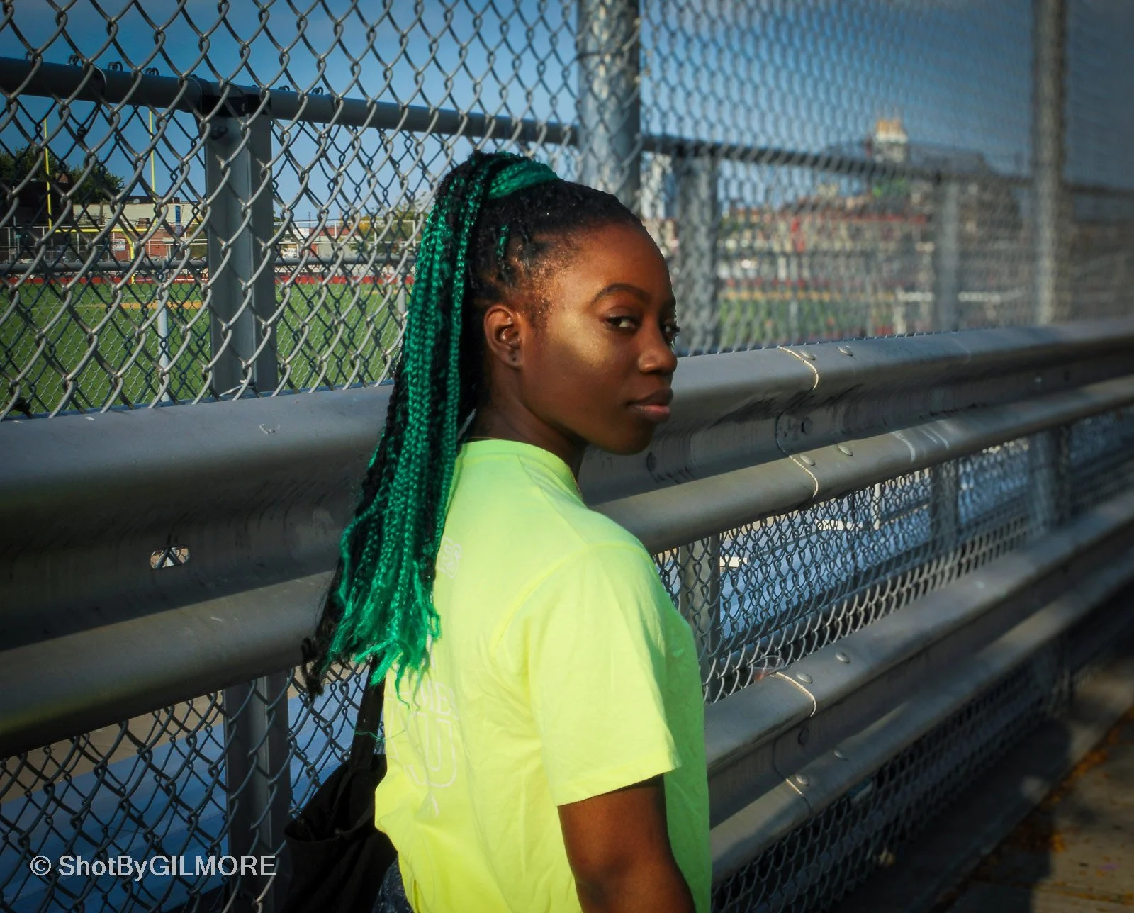 Young woman with green and black braided hair standing next to a chain-link fence at a sports field during daytime.