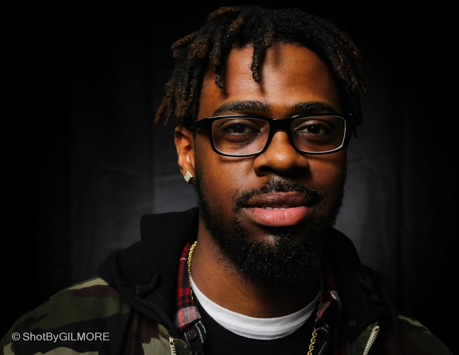 A portrait of a man with glasses, dreadlocks, a beard, and earrings, wearing a camouflage jacket and a gold chain, against a dark background.