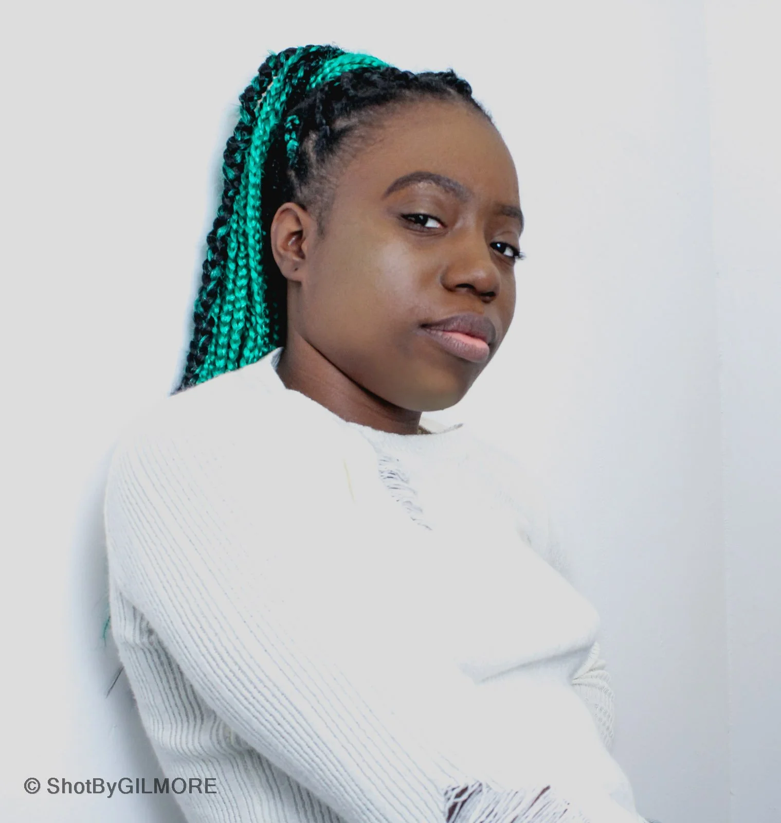 A young woman with braided hair with green tips, wearing a white distressed sweater, standing against a plain white background.