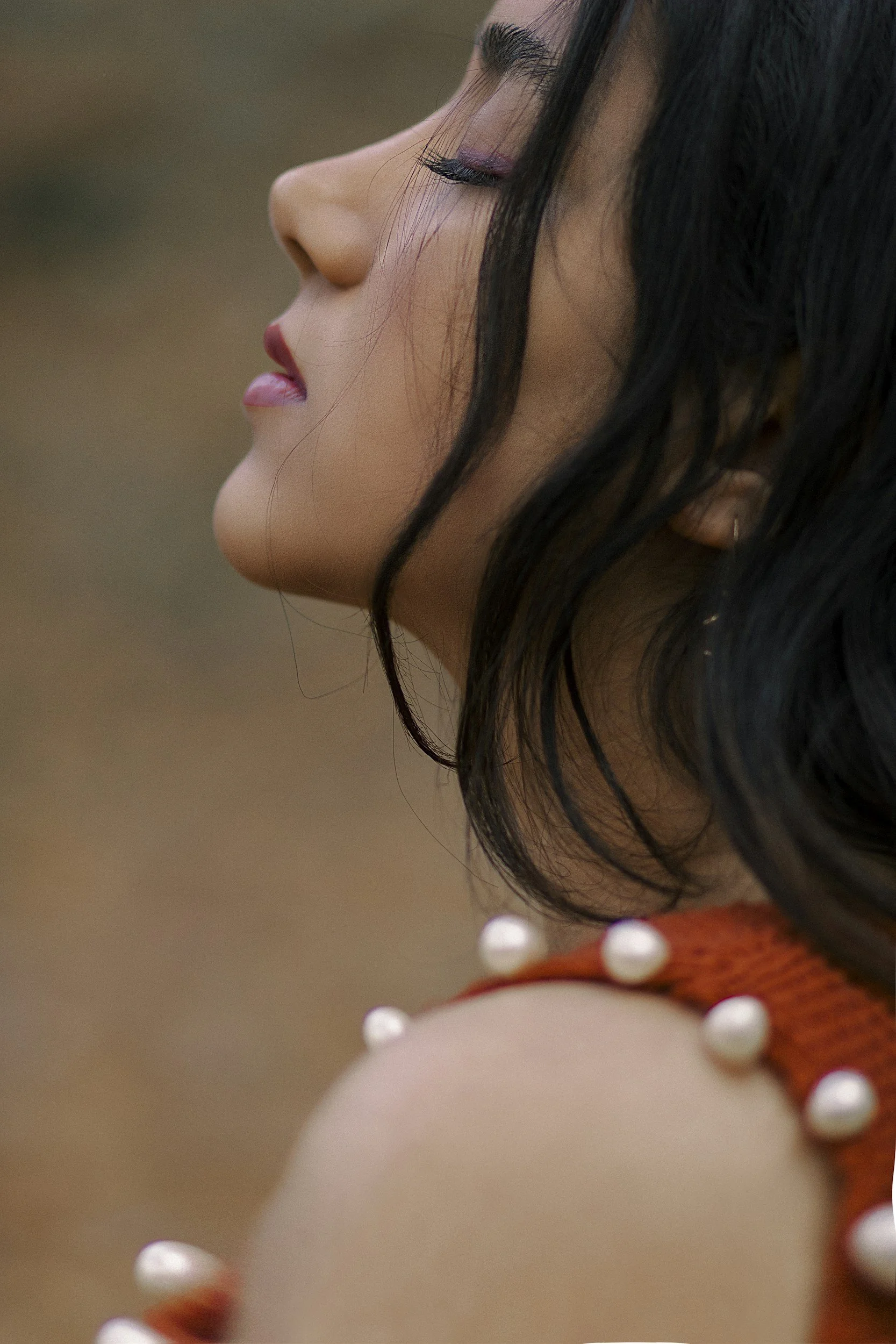 Close-up profile of a woman with closed eyes, black hair, and makeup, wearing an orange top with white pearls on the shoulder.