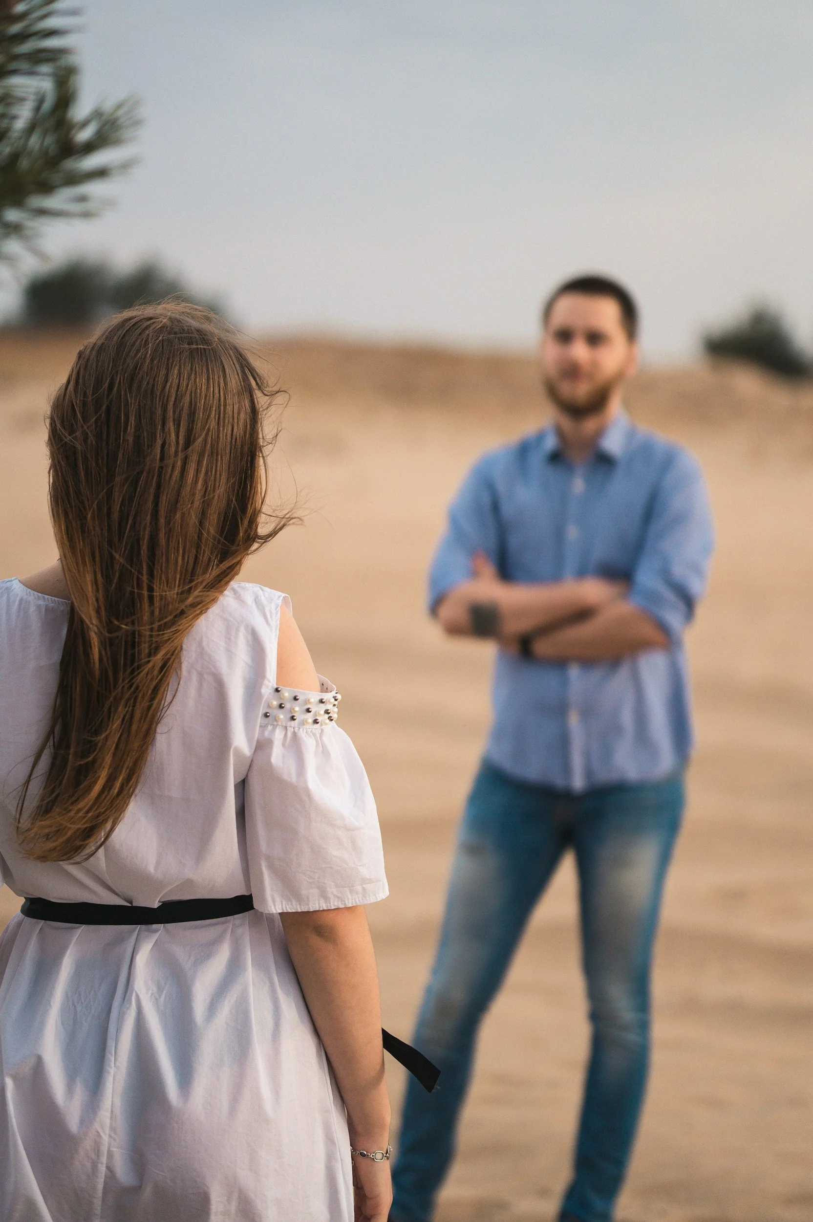 A woman with long hair wearing a white dress with shoulder embellishments and a black belt, standing outdoors, facing a man in a blue shirt and jeans with arms crossed, in a sandy or desert-like landscape.