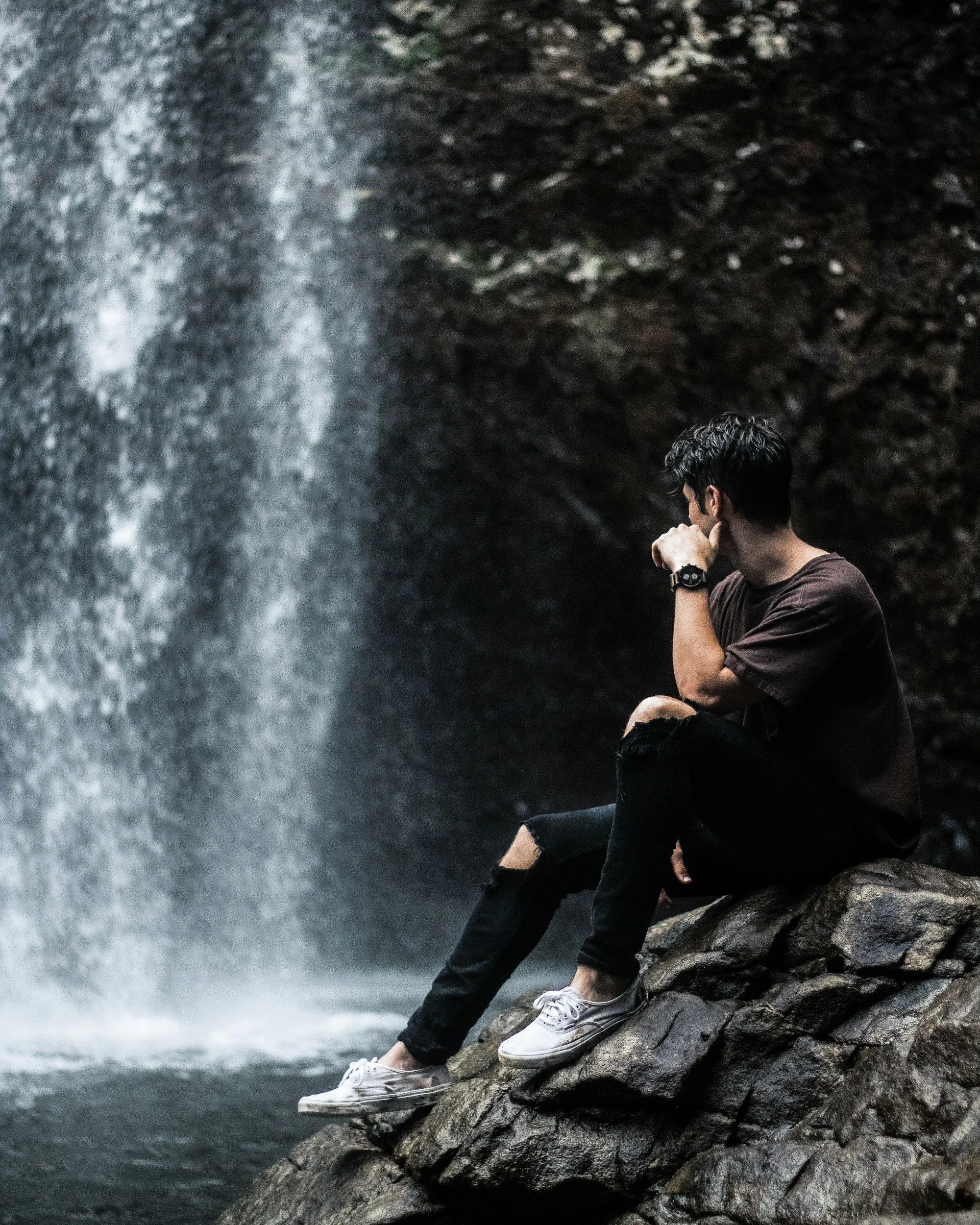 A young man sitting on rocks next to a waterfall, with his hand on his chin, looking at the waterfall.