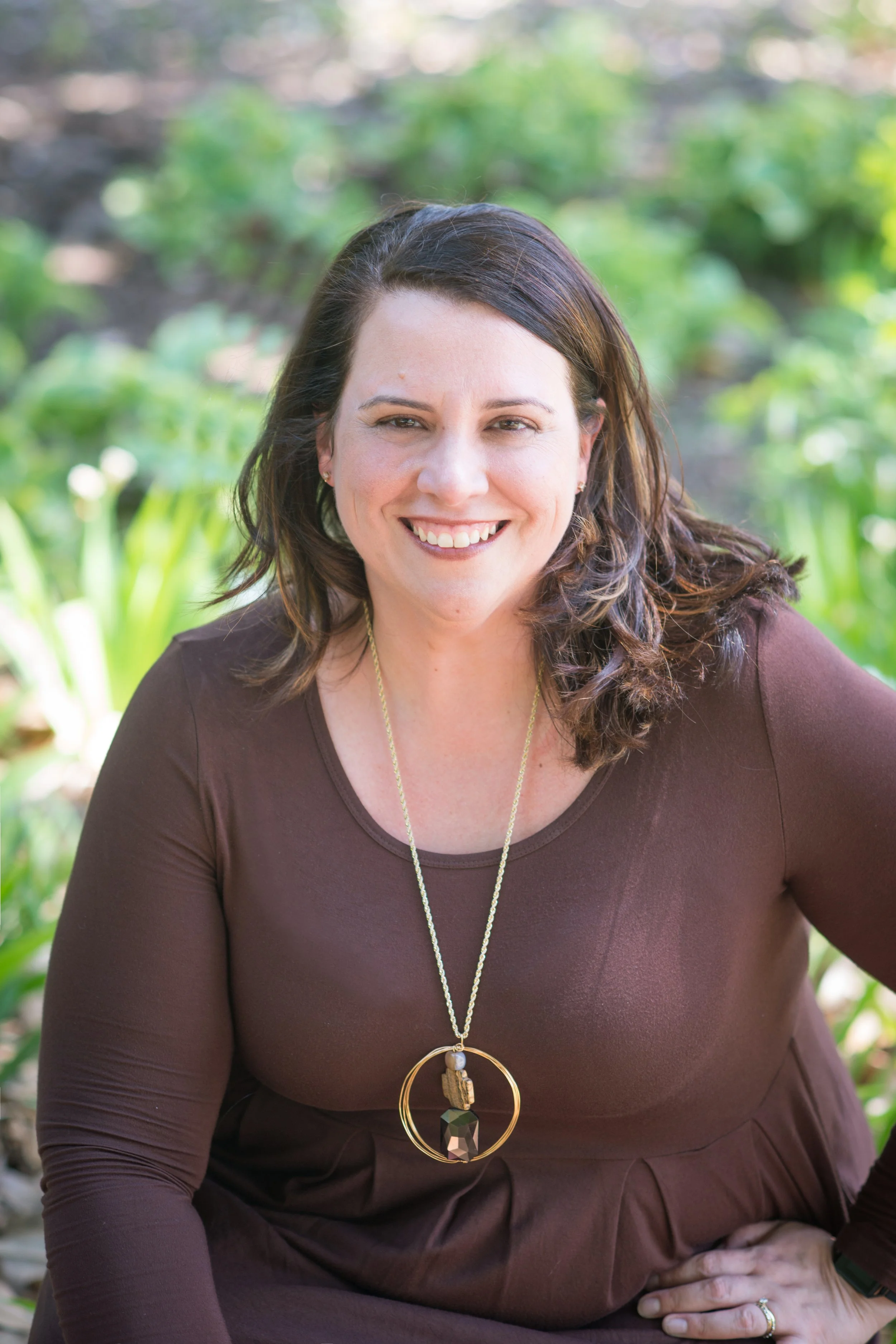 Woman smiling outdoors, wearing a dark brown dress and a long necklace, with greenery in the background.